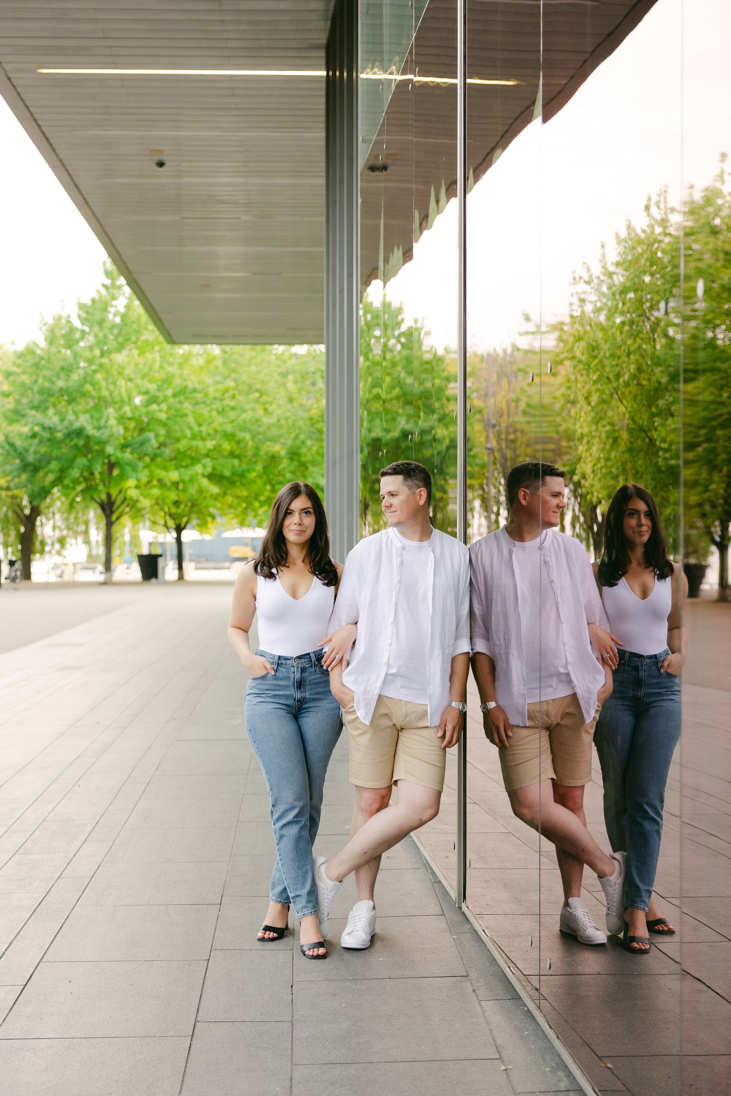 A couple poses against a reflective glass wall of a modern building in casual attire.