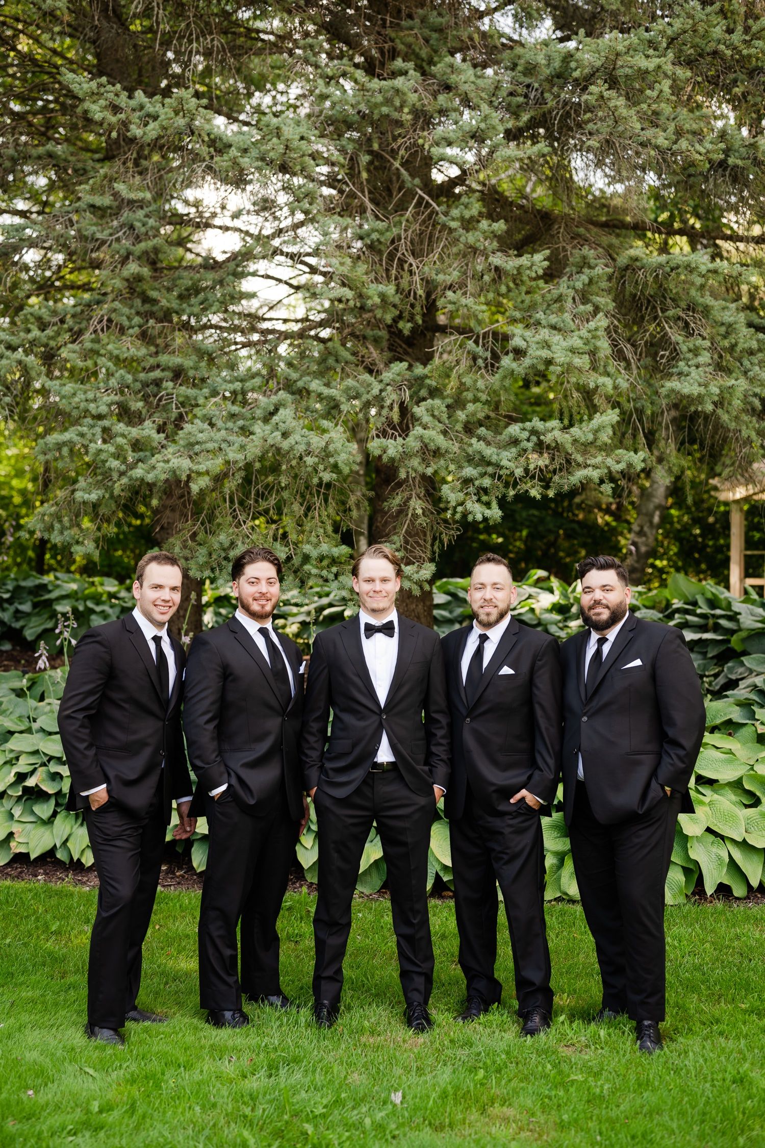 Five groomsmen in classic black tuxedos pose against a backdrop of lush garden foliage.
