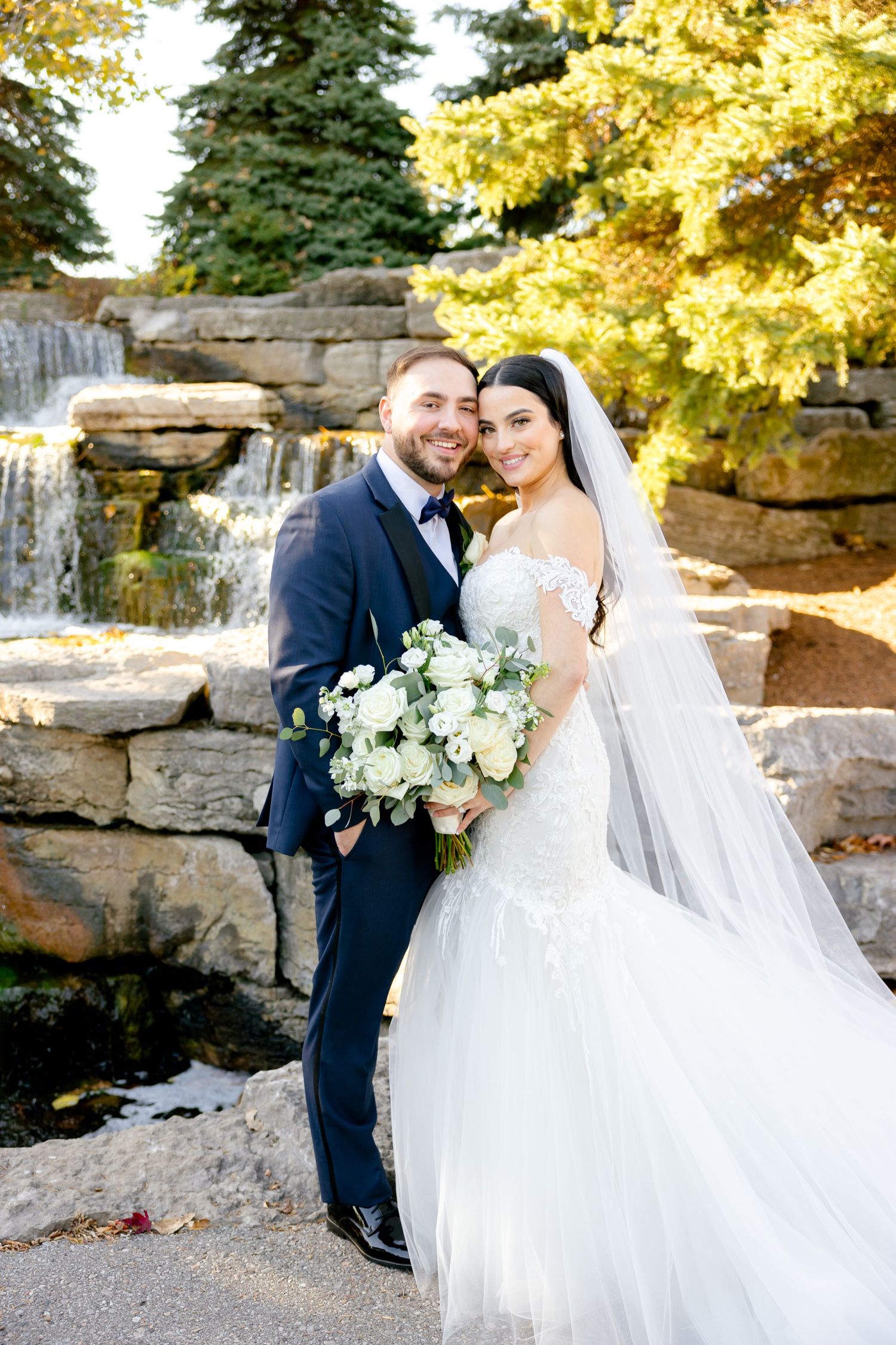 Wedding portrait in front of stone waterfall feature with autumn foliage in background.