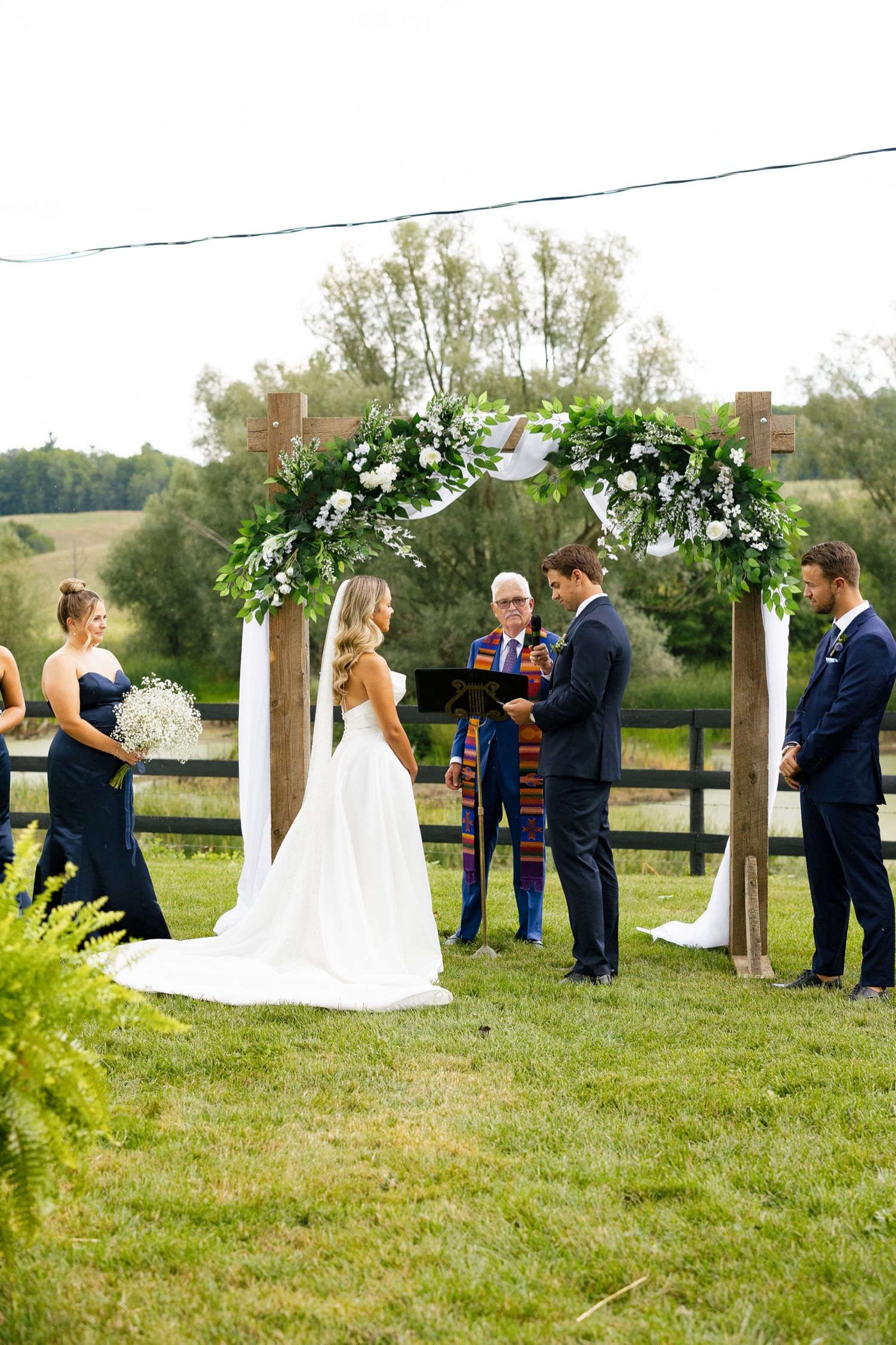 Wedding ceremony takes place beneath a floral-adorned wooden arch in a pastoral setting.
