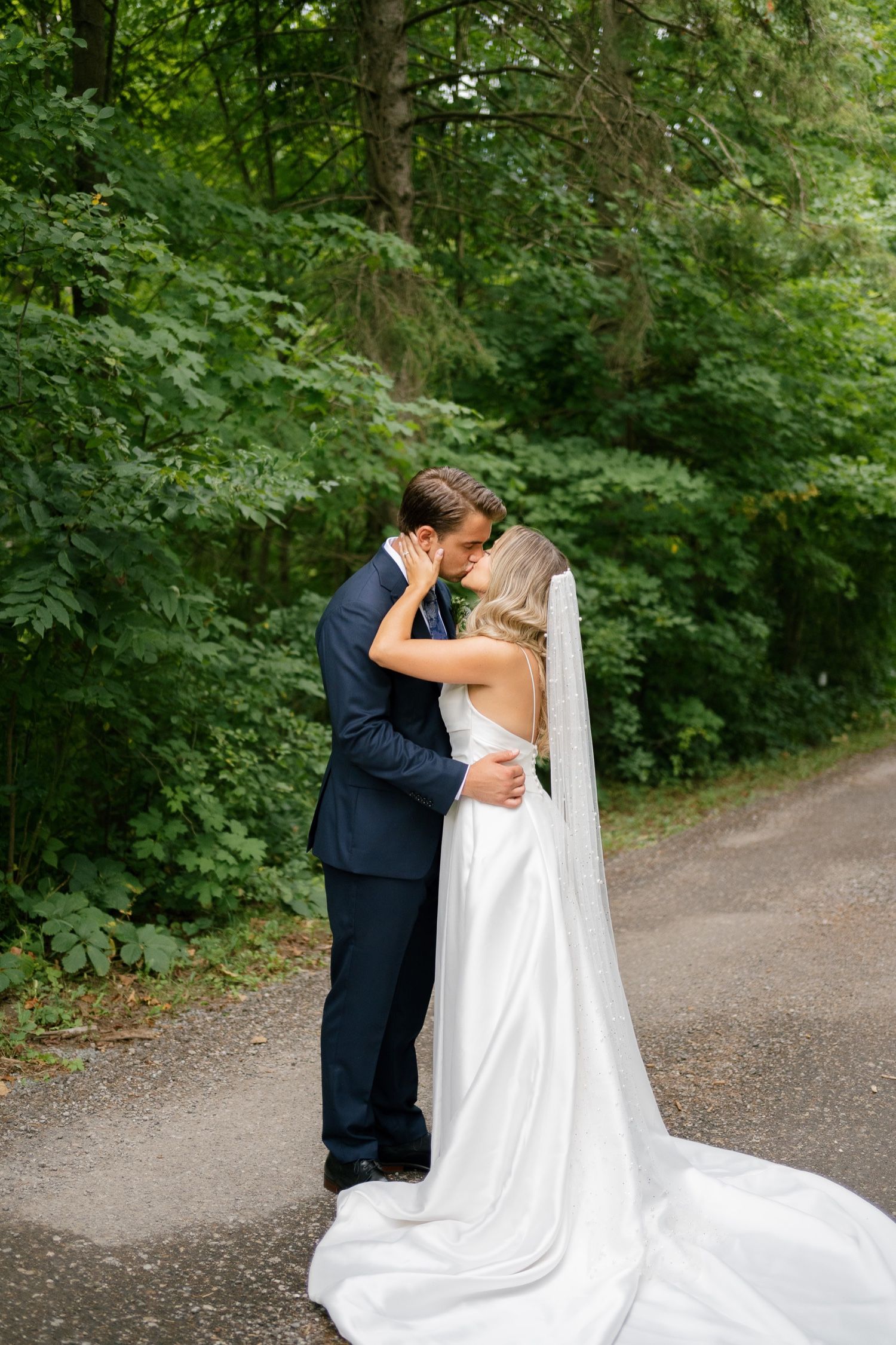 Tender embrace between newlyweds on forest path with dramatic wedding dress train.