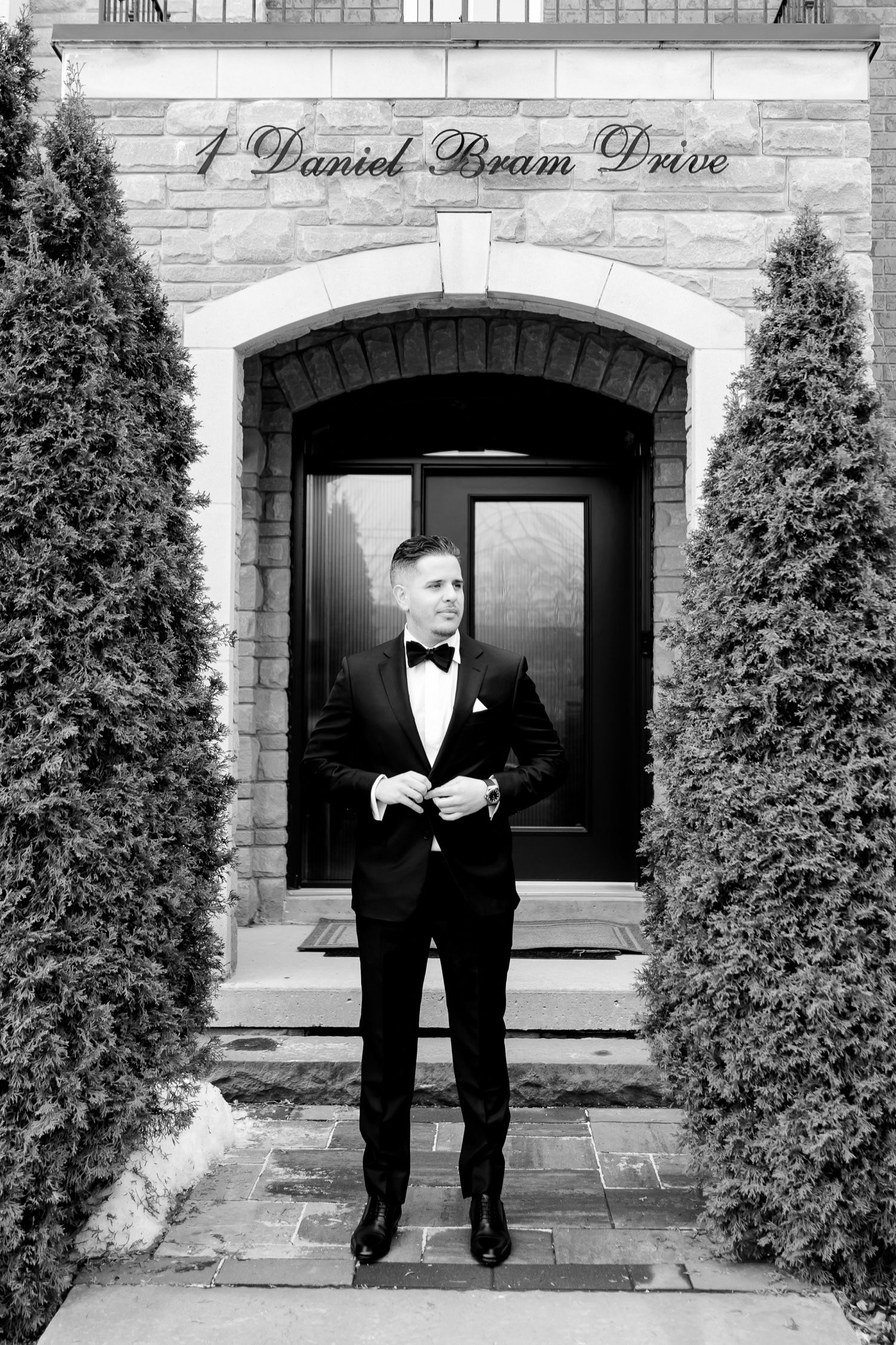 Black and white portrait of groom at elegant stone entrance with cypress trees.