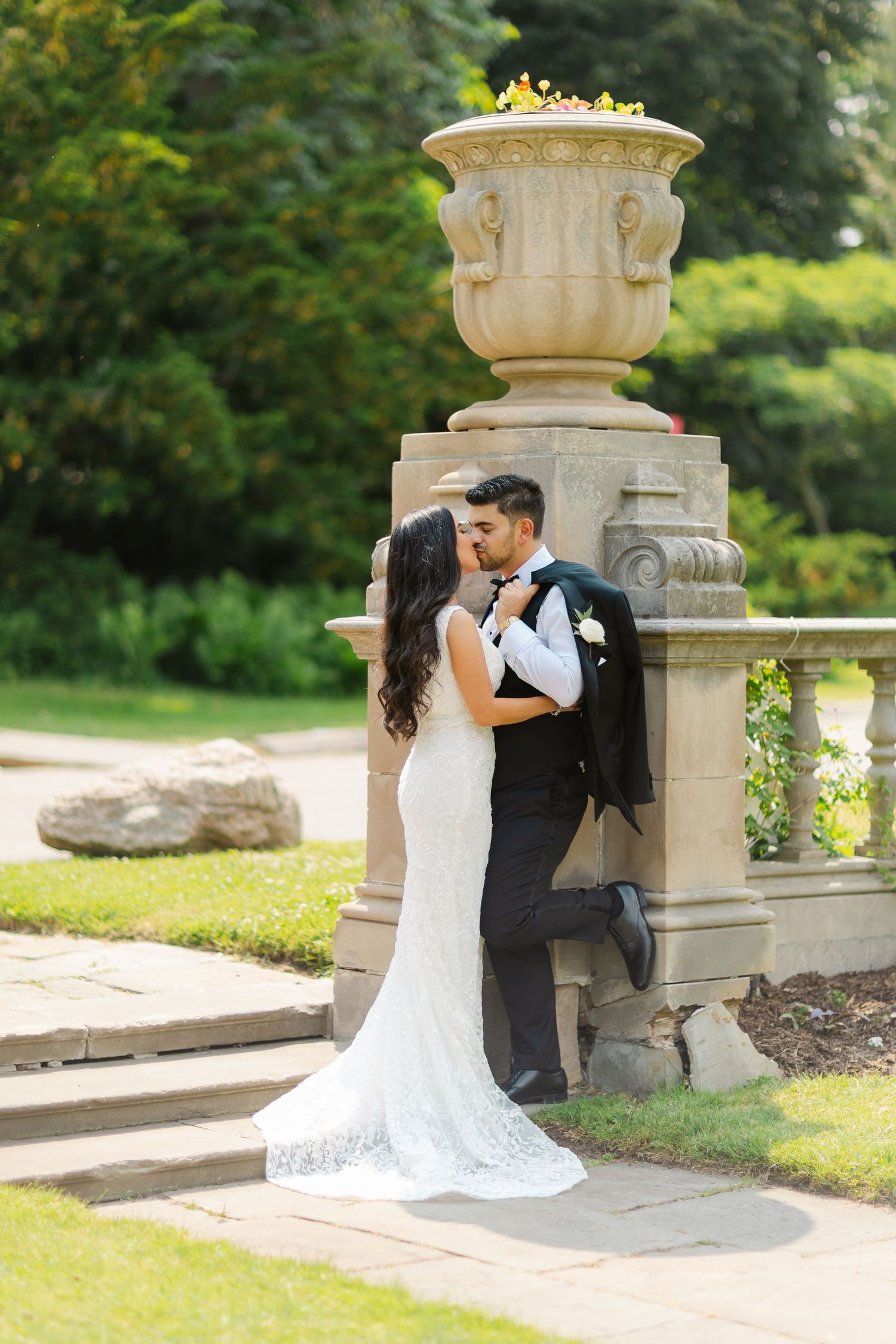 A romantic wedding couple embraces by an ornate stone urn pillar in a garden setting.