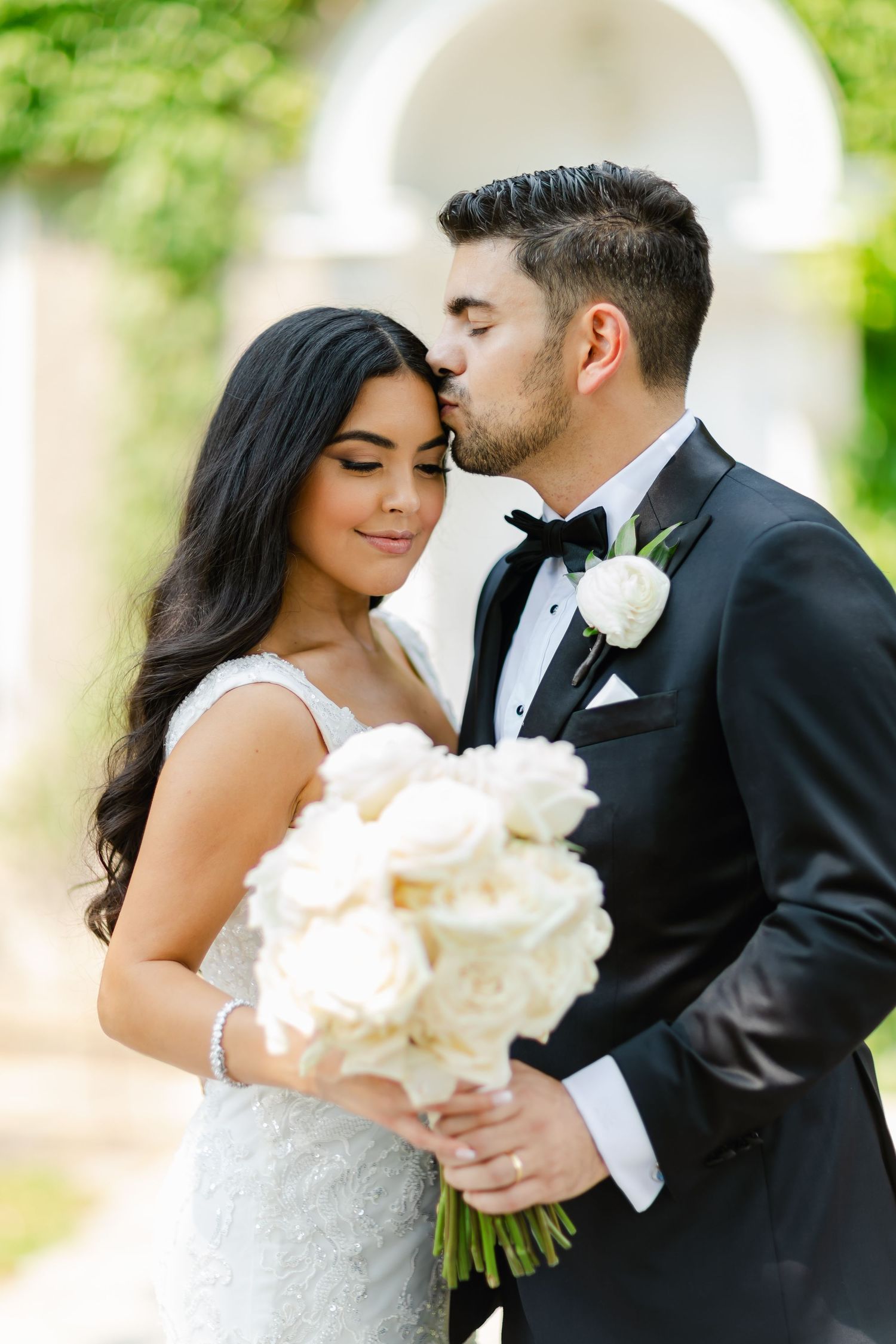 Romantic wedding portrait with couple embracing under white arch with bridal bouquet.