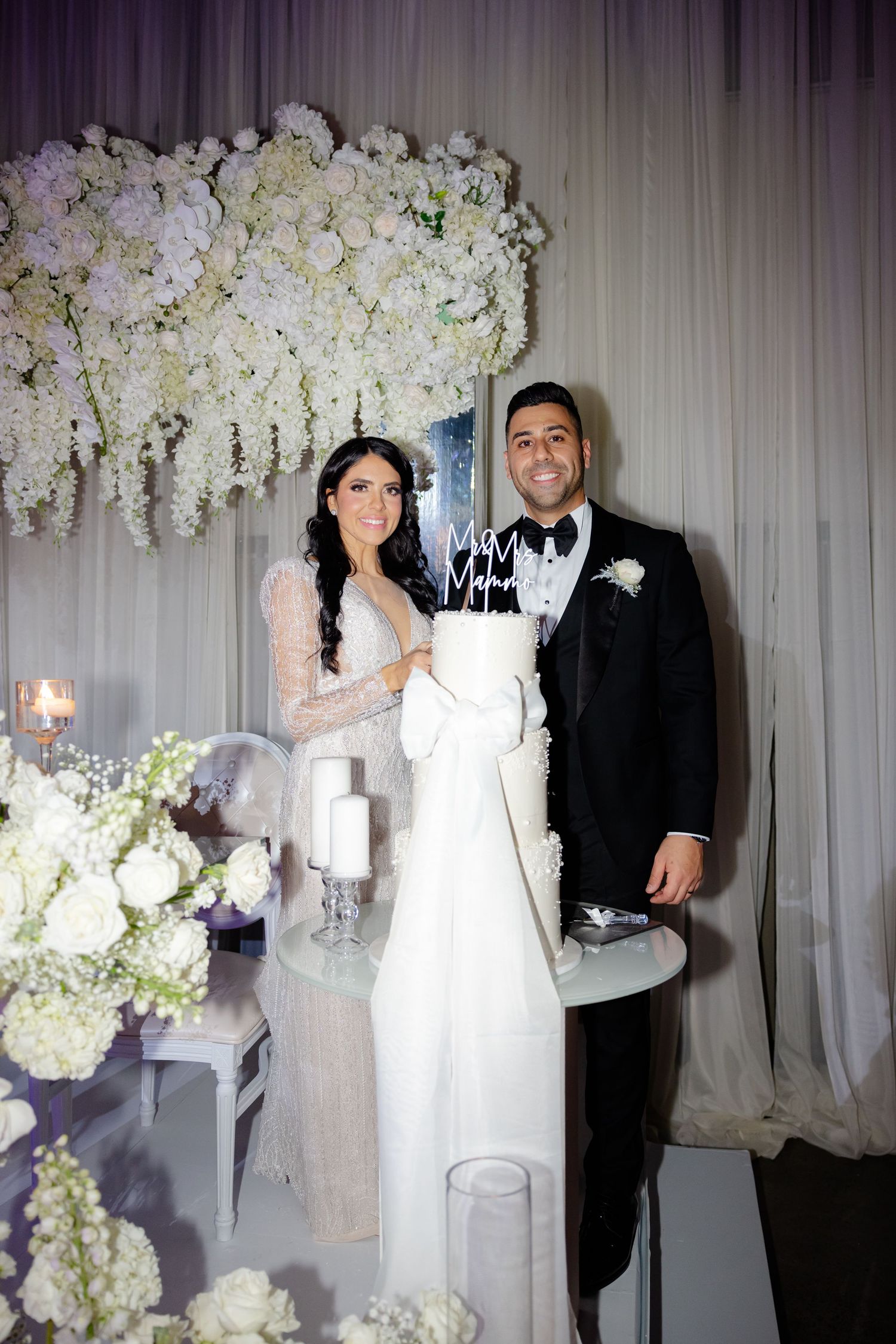 Newlyweds pose with their wedding cake surrounded by elegant white floral decorations.