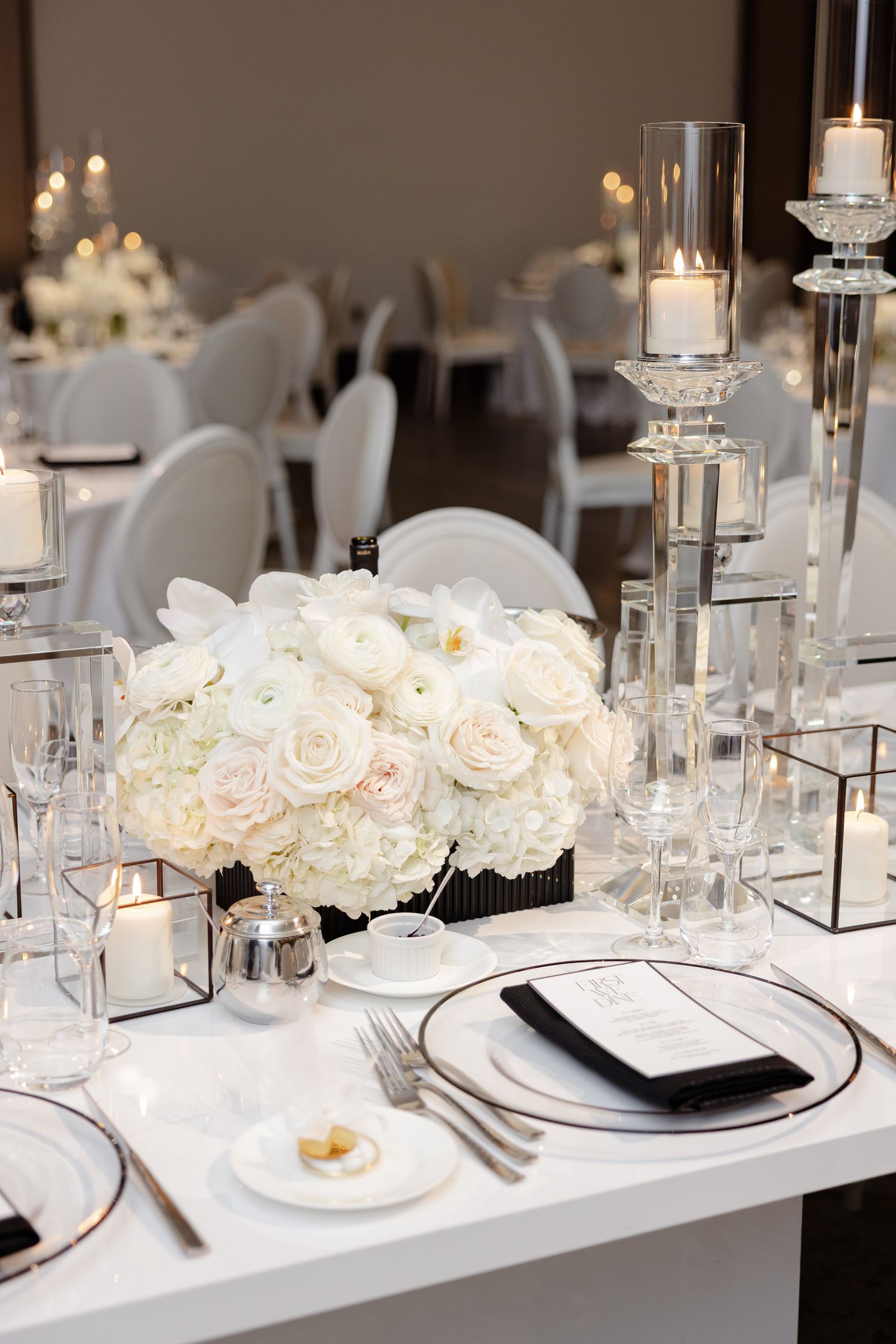 Modern white table setting with crystal candle holders and lush white rose centerpiece at formal wedding reception.