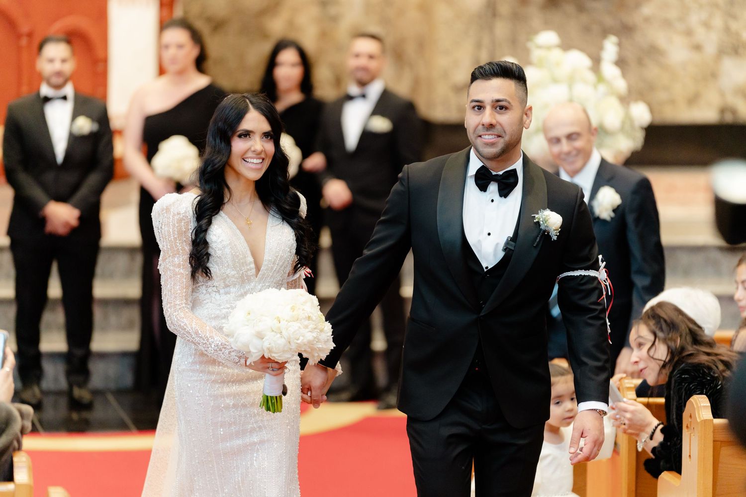 Newlyweds walk down the red-carpeted church aisle with wedding party and guests in the background.