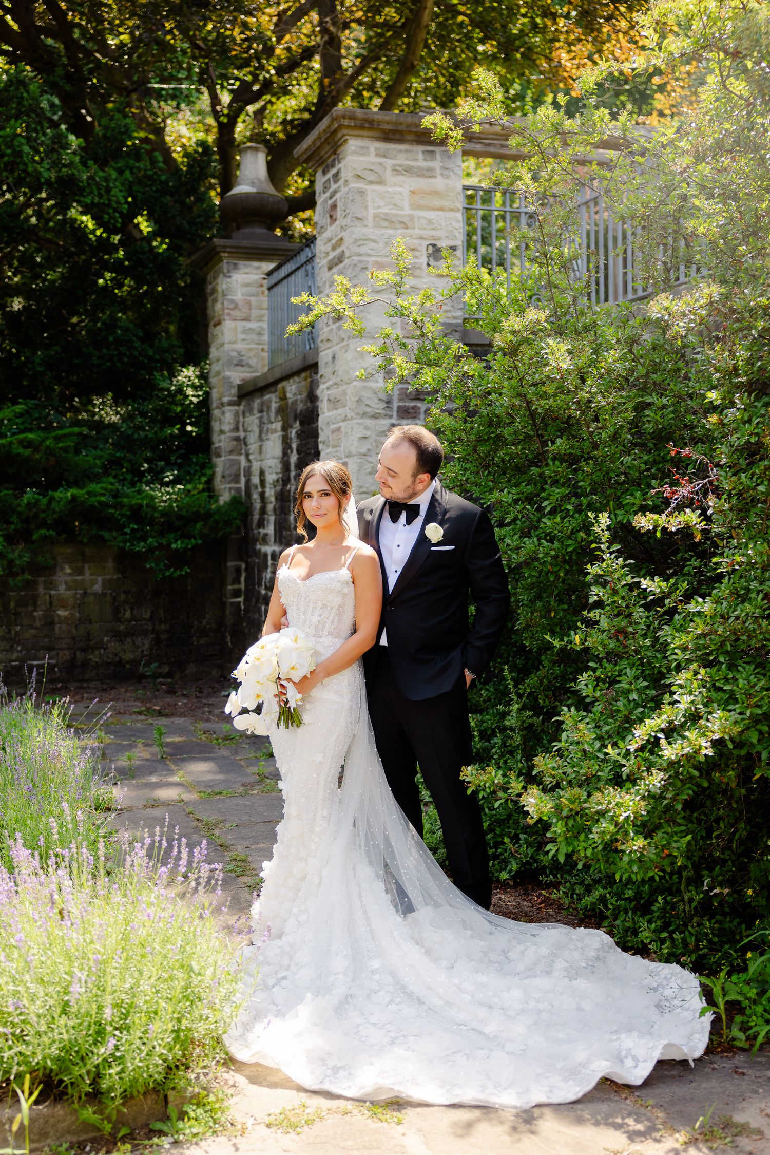 A newlywed couple poses in formal attire by a stone wall and garden gate on their wedding day.