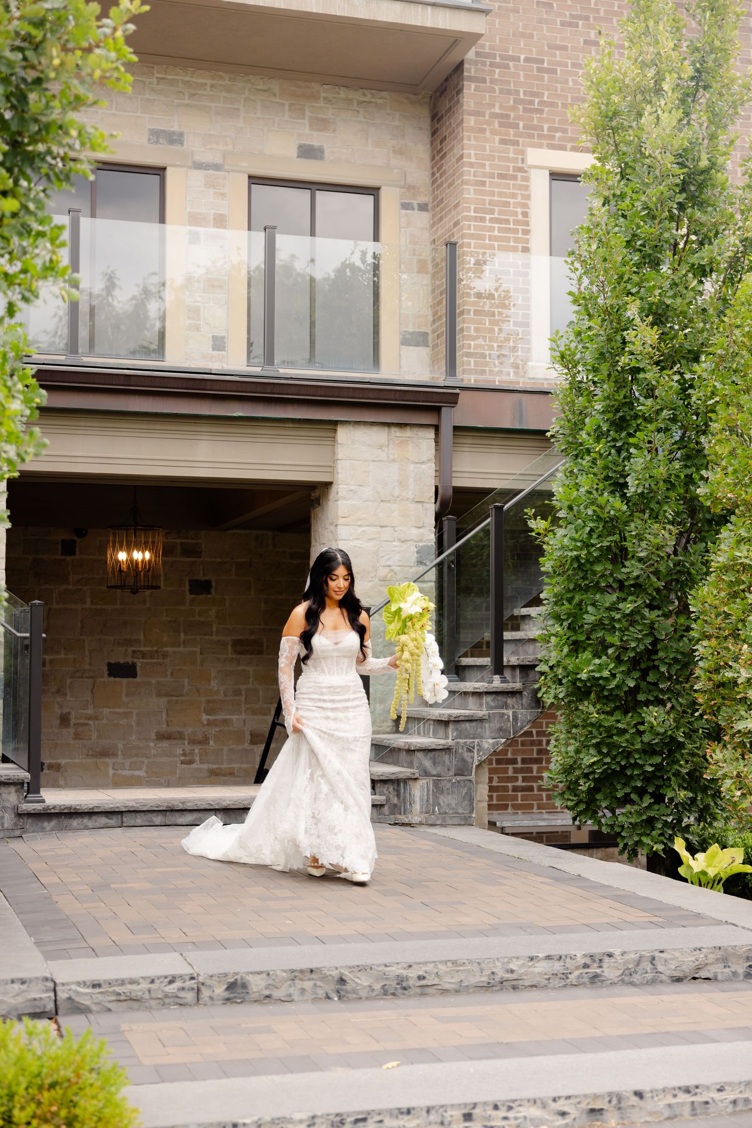 A bride in an elegant white lace wedding gown descends stone steps outside a modern brick building with greenery.