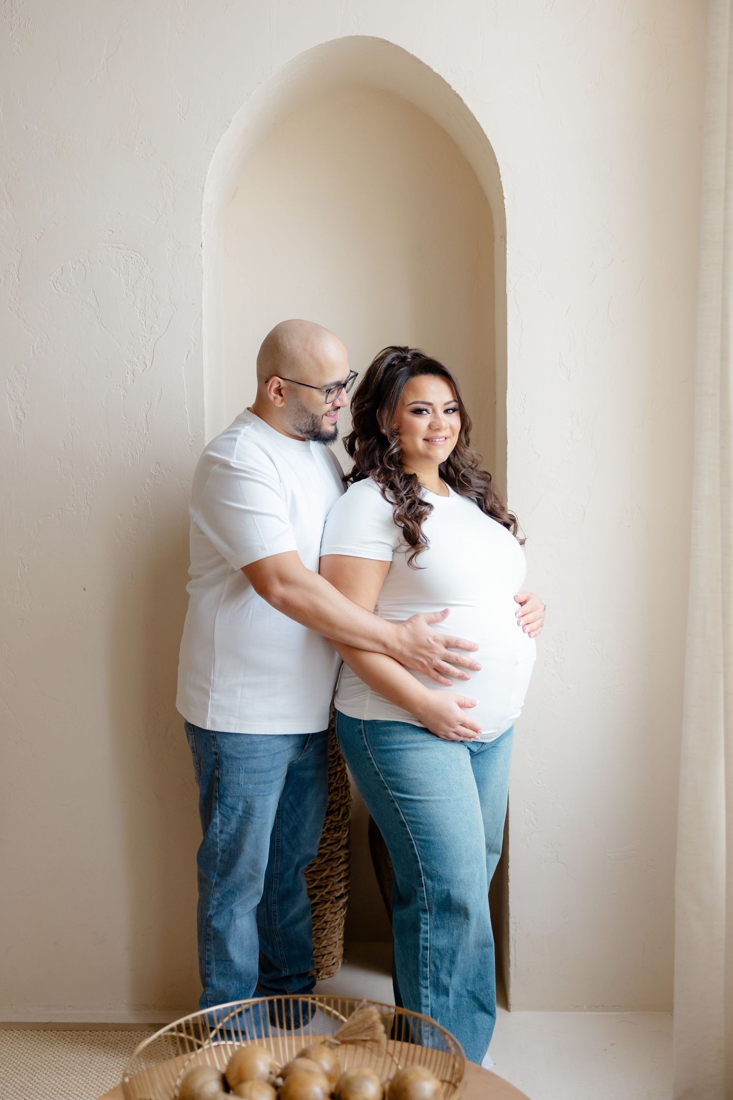 A couple in white shirts and jeans stand in an arched doorway during a maternity photoshoot.