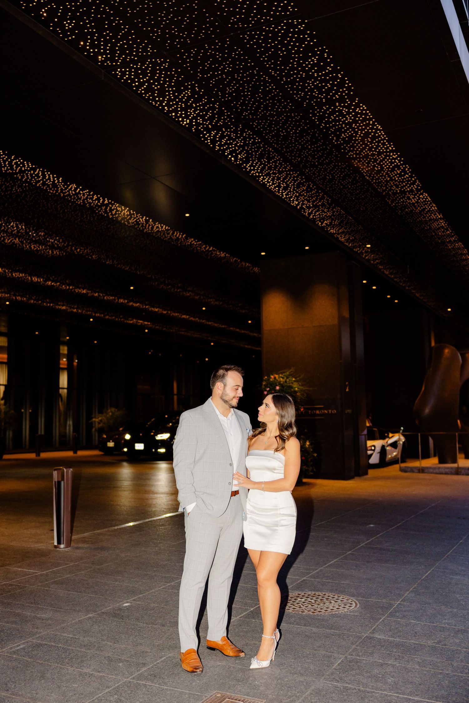 Couple shares romantic moment under sparkly lights in urban setting.