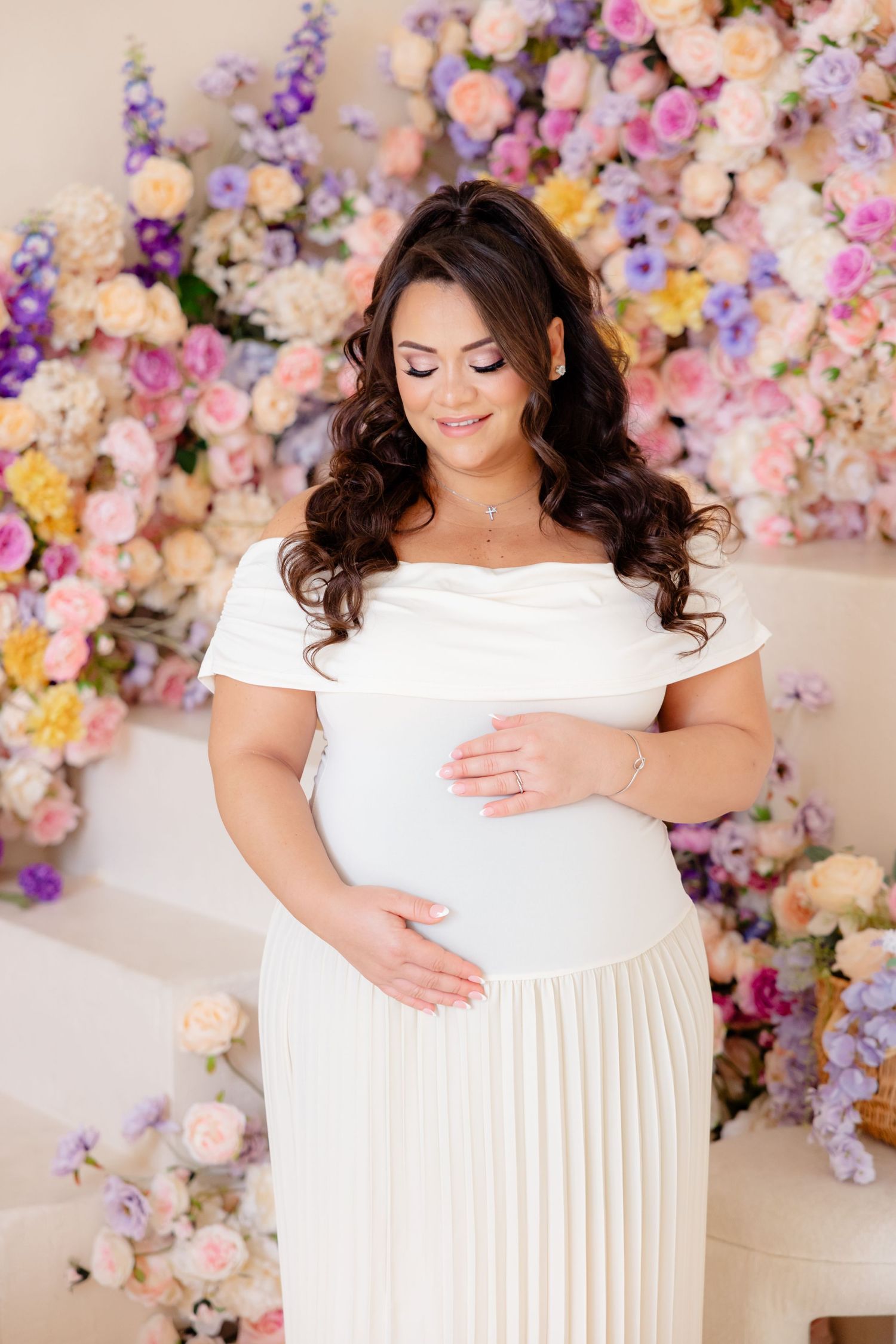 Pregnant woman in white dress poses against floral backdrop with pink and purple flowers for maternity photoshoot.