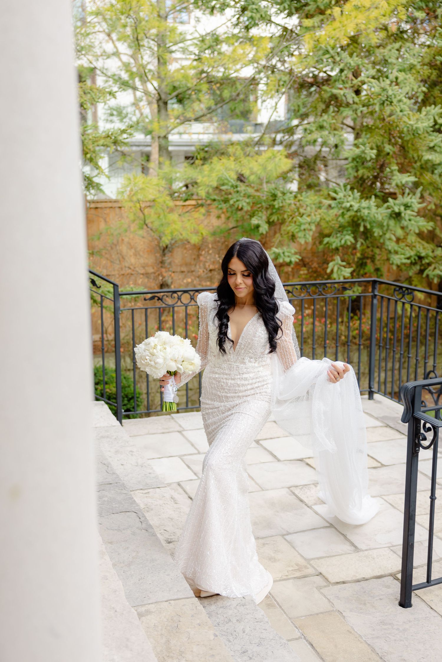 Bride in fitted lace gown holds bouquet while posing on stone balcony with garden backdrop.