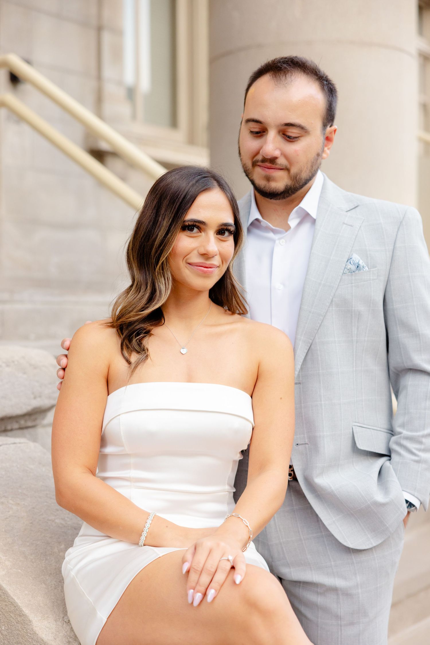 Couple in formal attire pose together on stone steps with the person in white dress seated and grey suit standing.