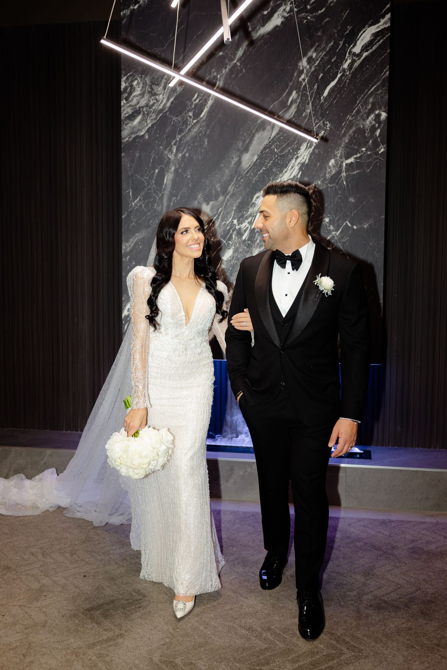 Newlyweds walking together in formal attire with bride holding white bouquet against dark marble background.