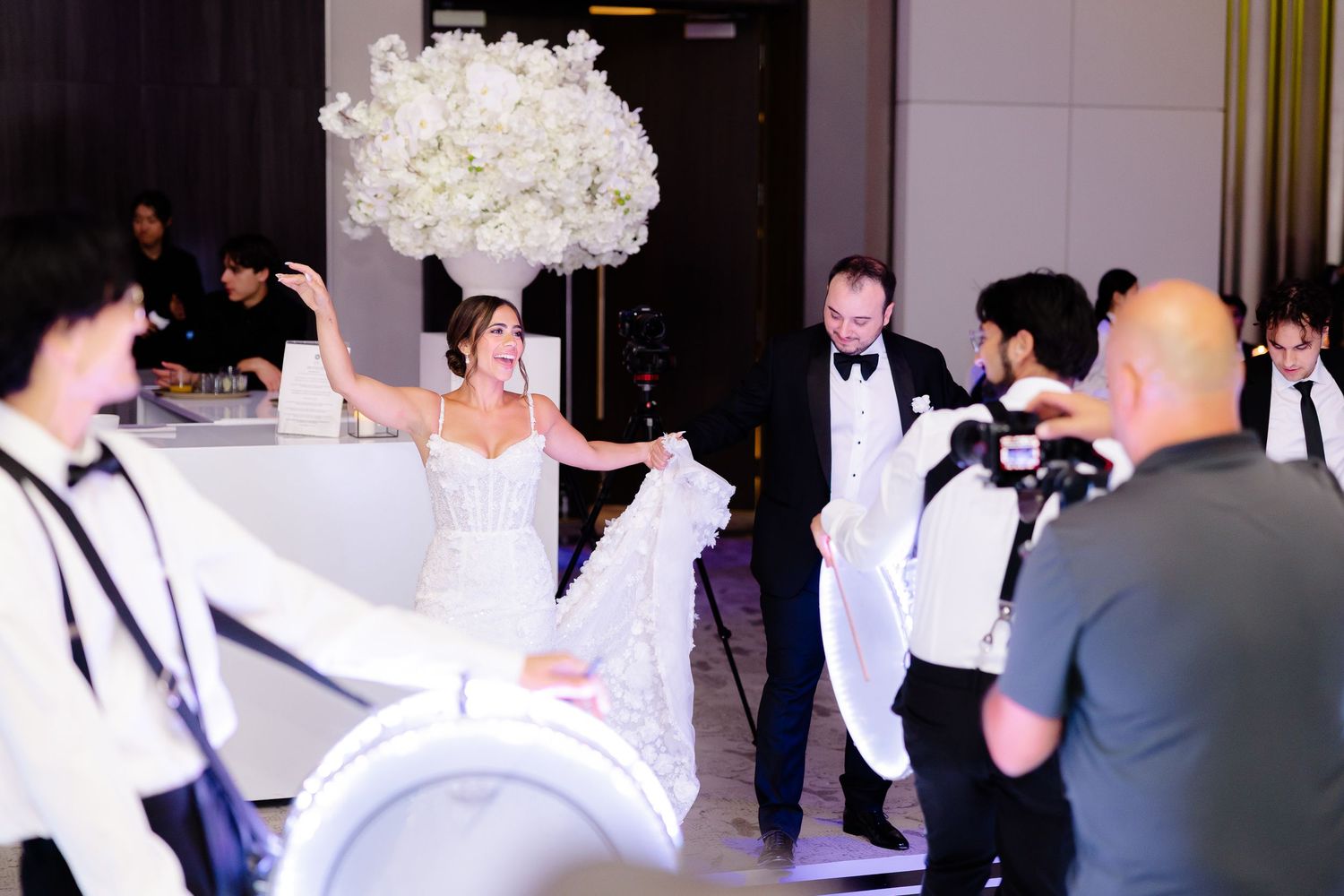 Guests in formal attire dance and celebrate at an elegant indoor wedding reception in white decor.