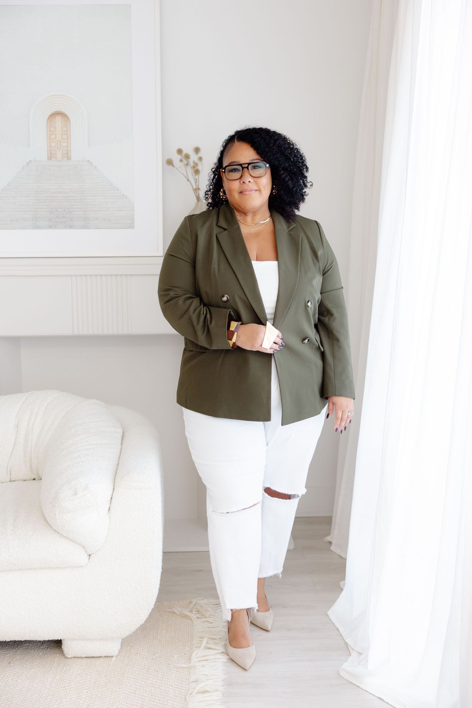 Professional outfit featuring an olive blazer with white tank top, distressed white jeans and neutral shoes in a bright setting.