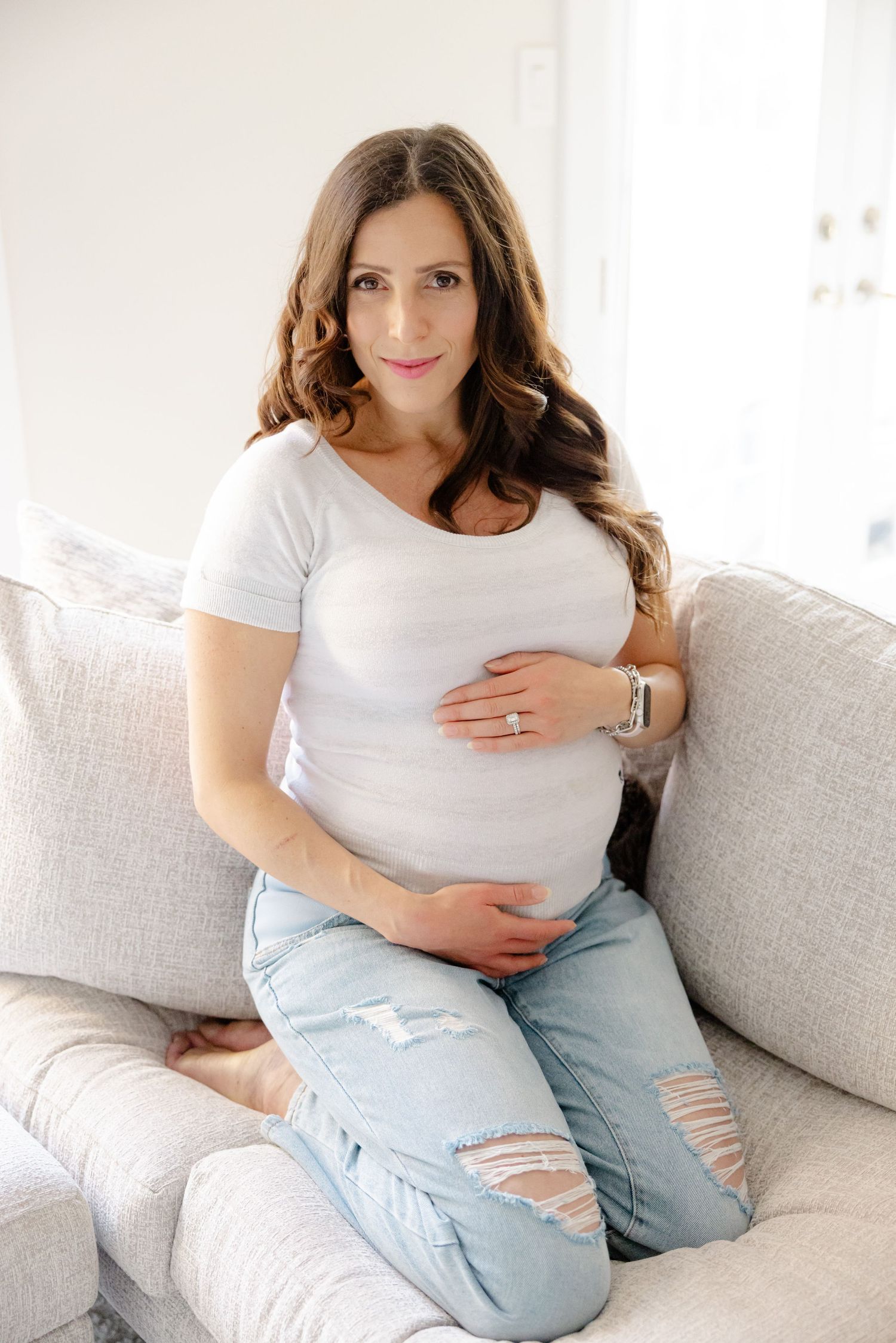 Expectant mother in white t-shirt and distressed jeans sits comfortably on light gray couch while cradling baby bump.