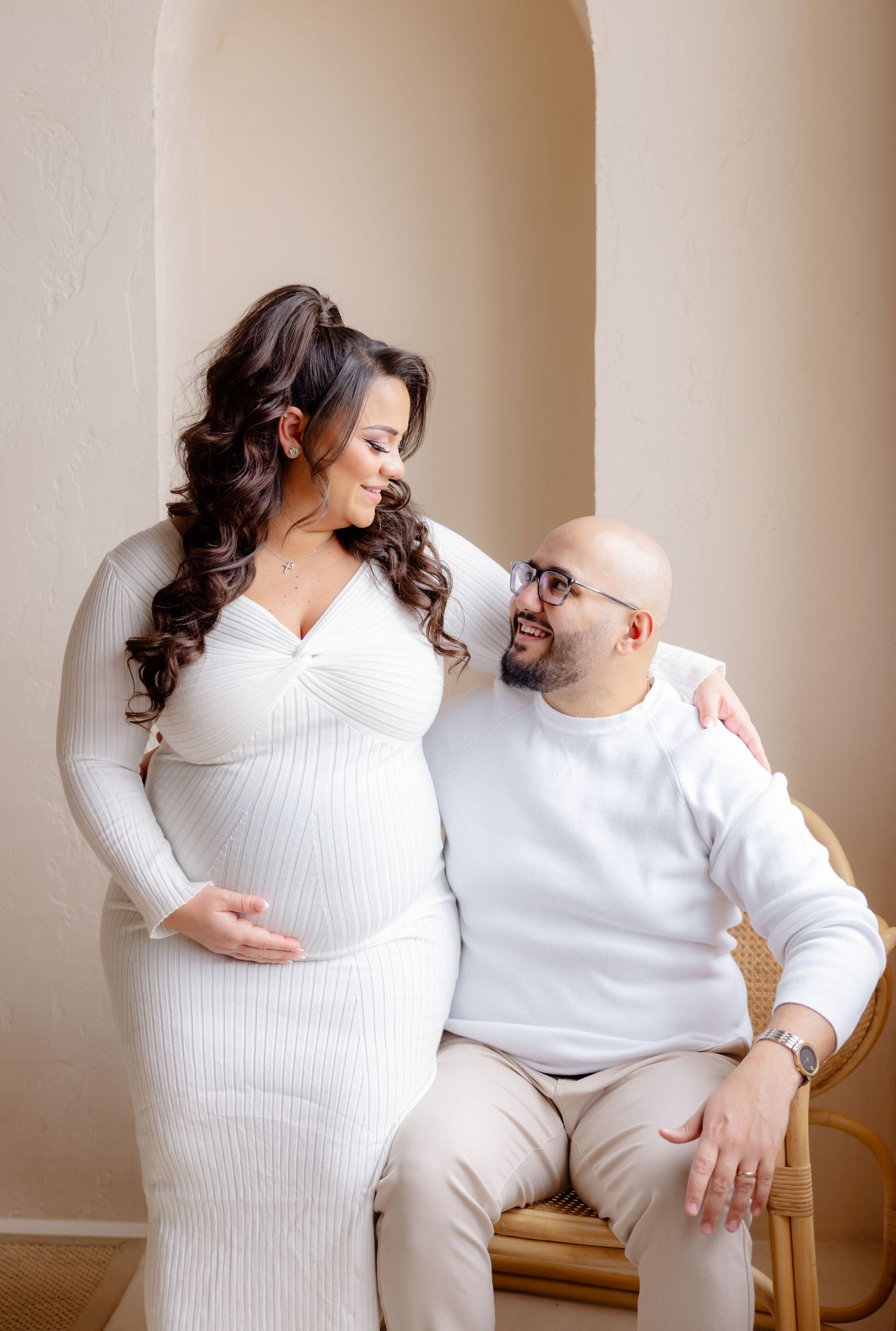 A romantic maternity photo of a couple in matching white sweaters smiling at each other against a neutral wall.
