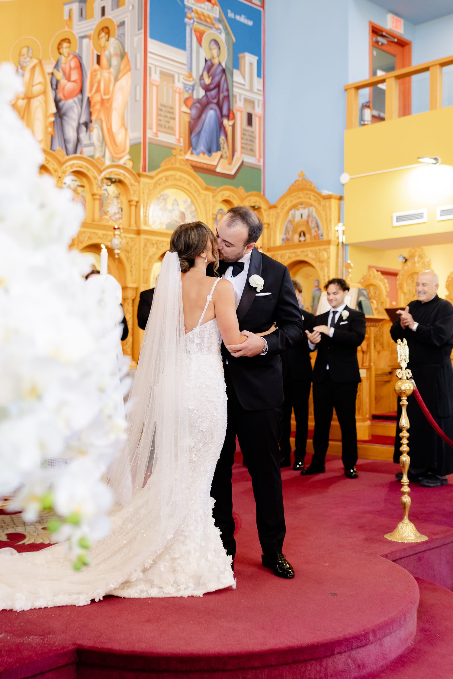 A couple shares their first kiss during a wedding ceremony in an ornate church with colorful religious murals and red carpeting.