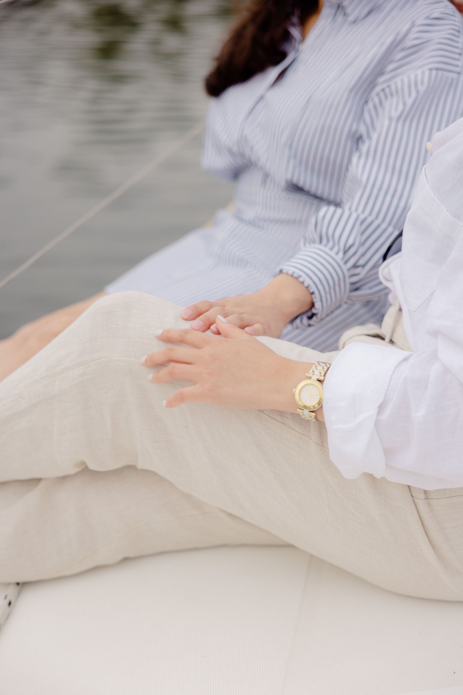 Close-up of hands with a gold watch resting on white-clad legs against a blurred background.