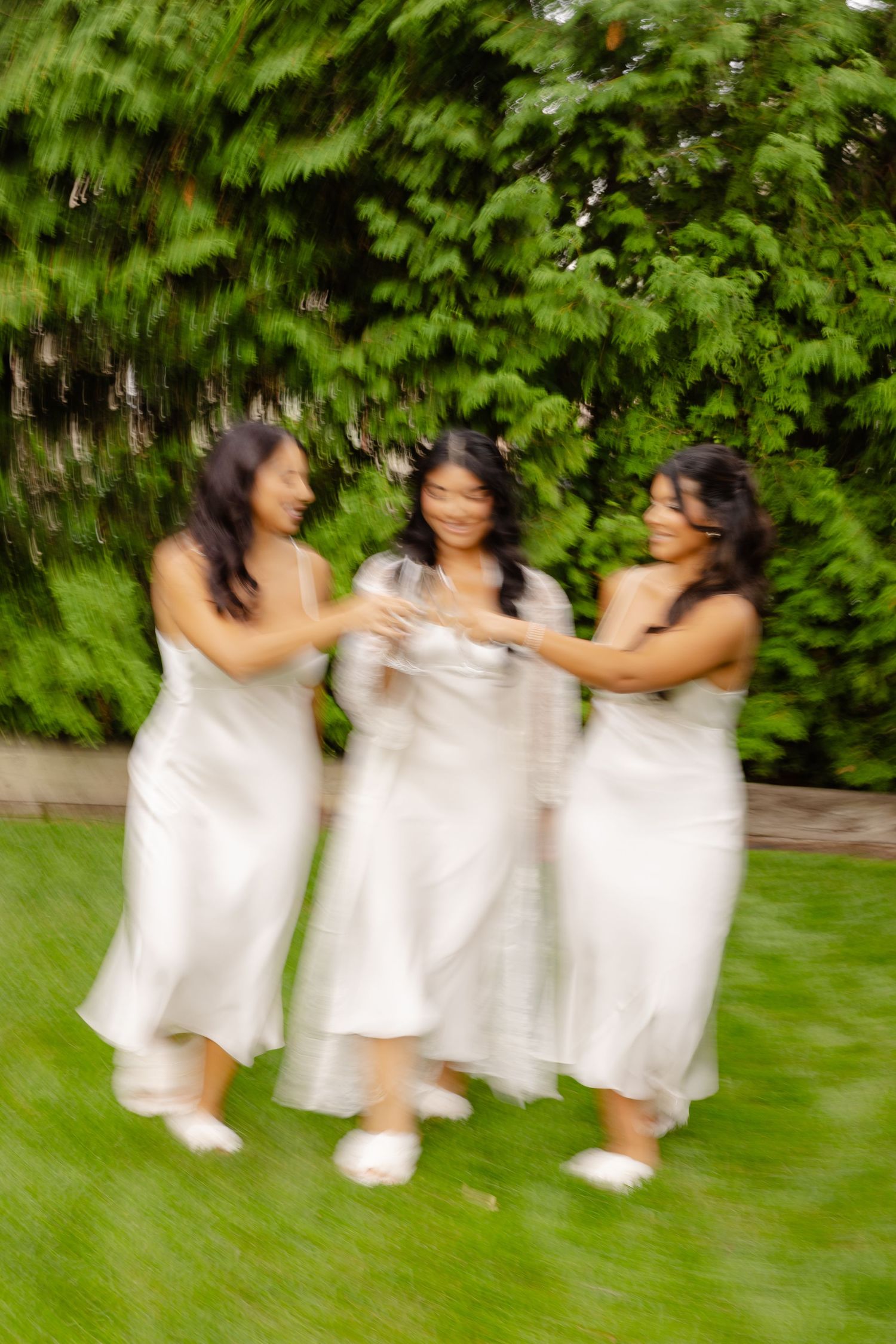 Three figures in flowing white dresses dance and twirl together on a green lawn against a leafy garden backdrop.
