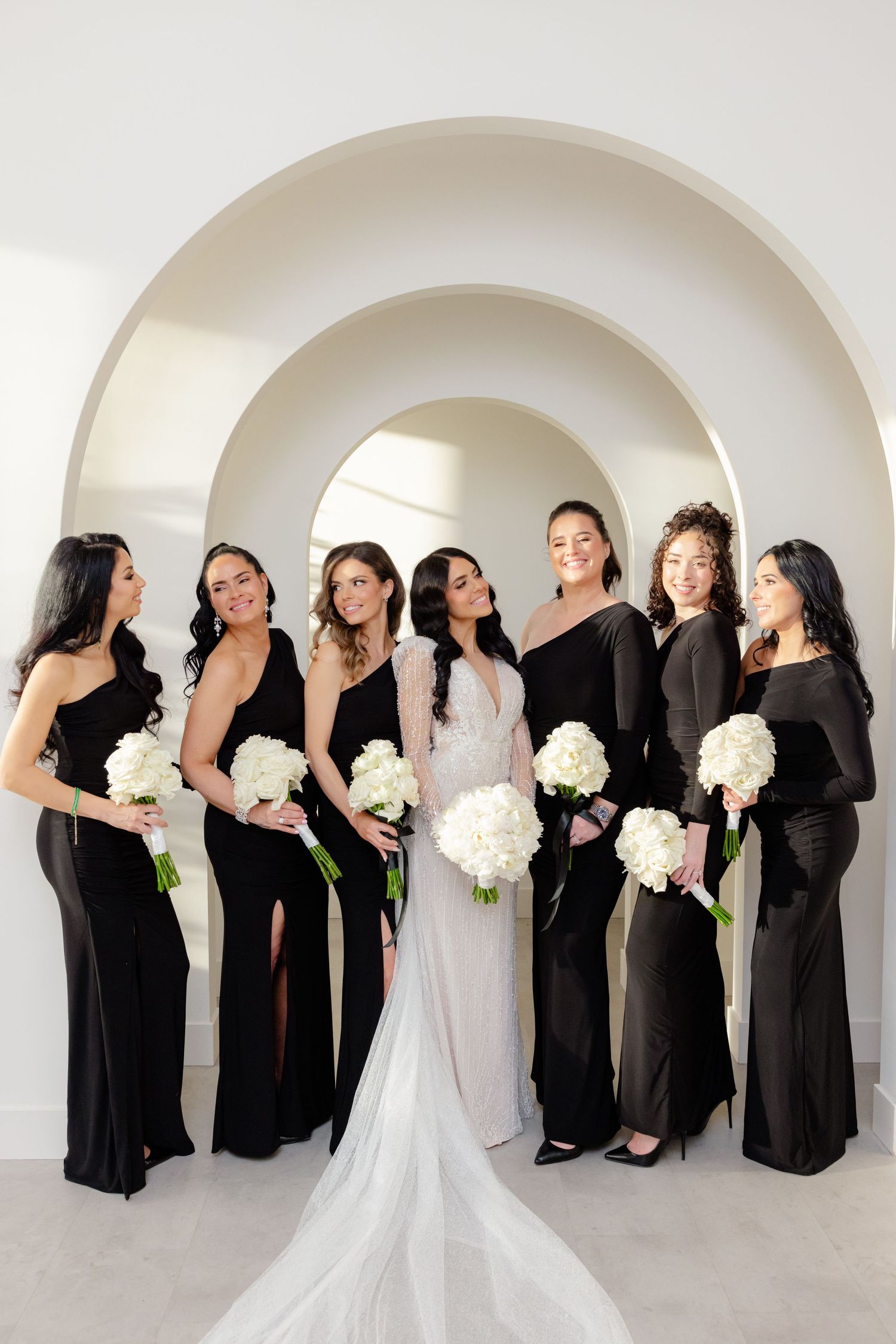 Bridal party in black gowns with white bouquets posed under modern nested arches.