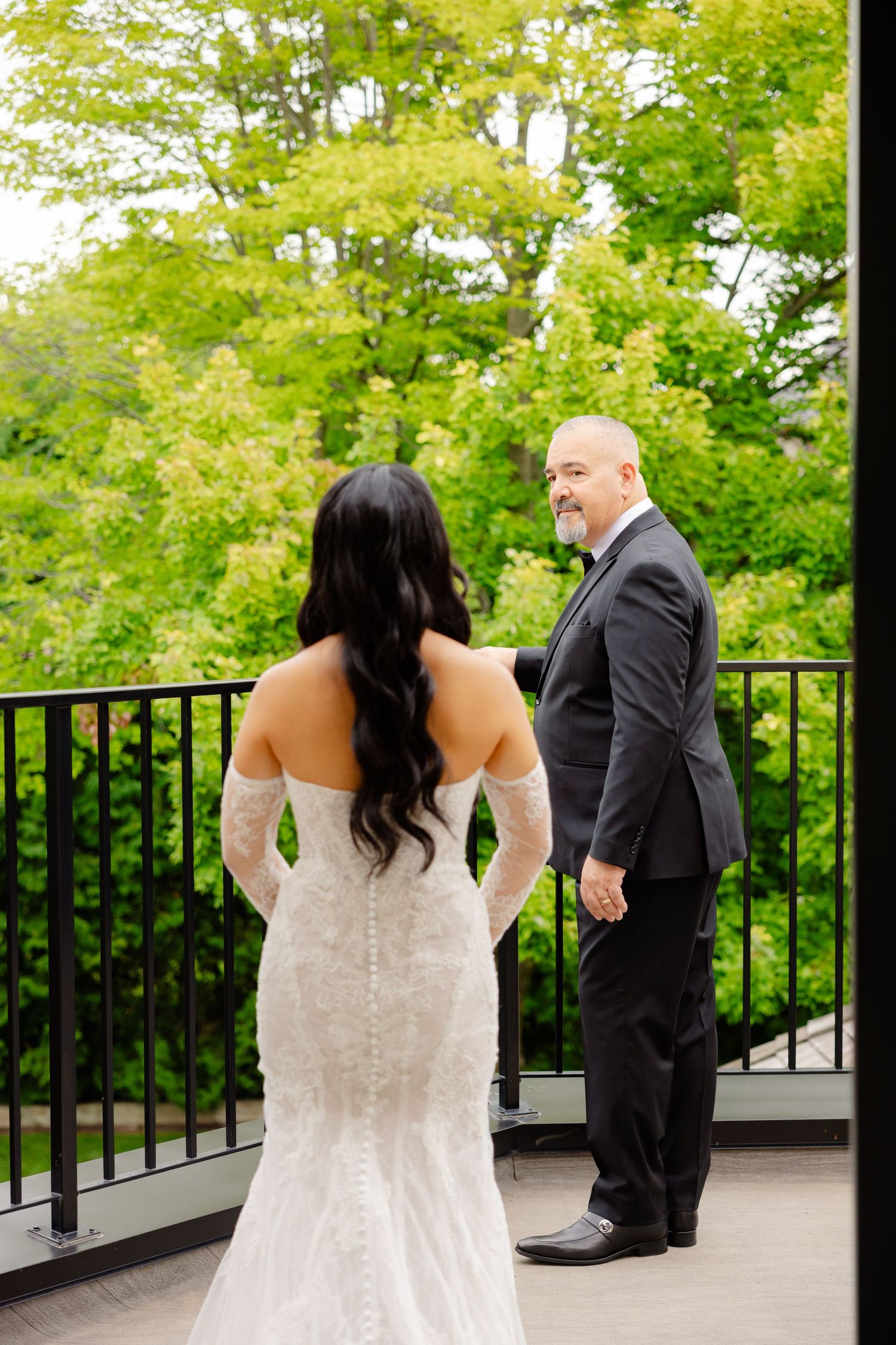 Couple shares first look moment on an outdoor deck surrounded by lush green trees on their wedding day.