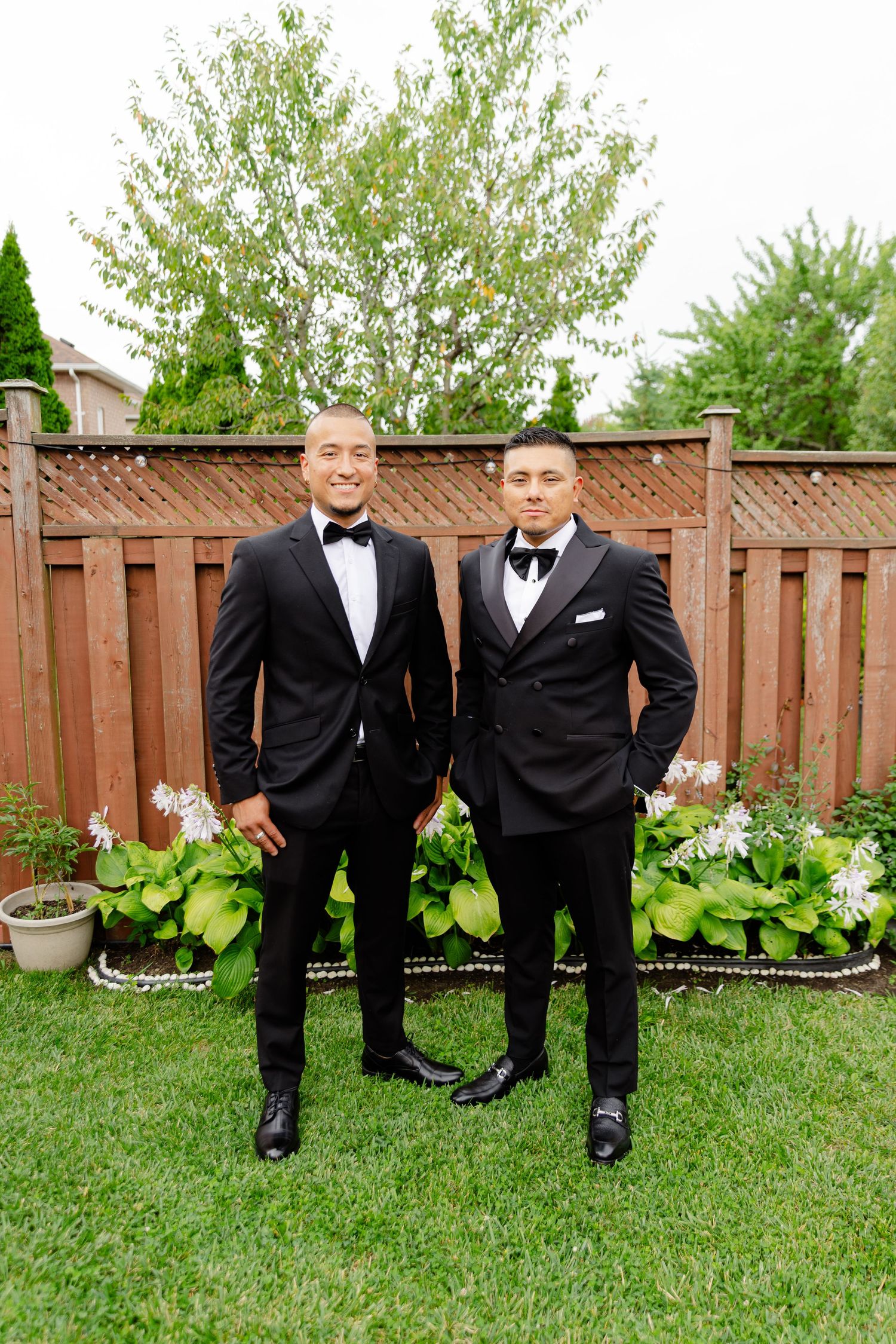 Groomsmen in black tuxedos and bowties pose together on grass lawn with wooden fence and flowers in background.