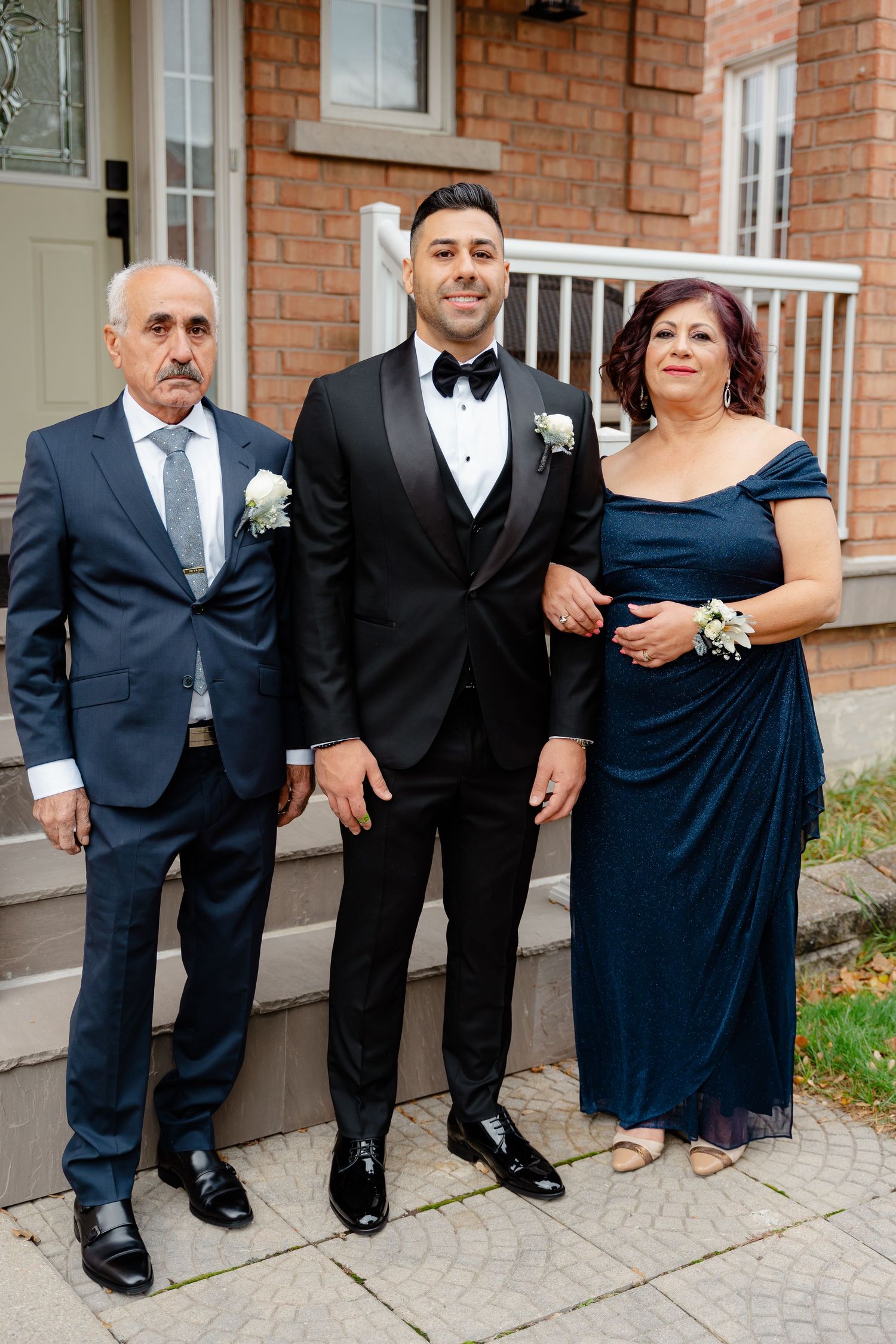 Three people in formal attire pose on brick house steps before a wedding celebration.
