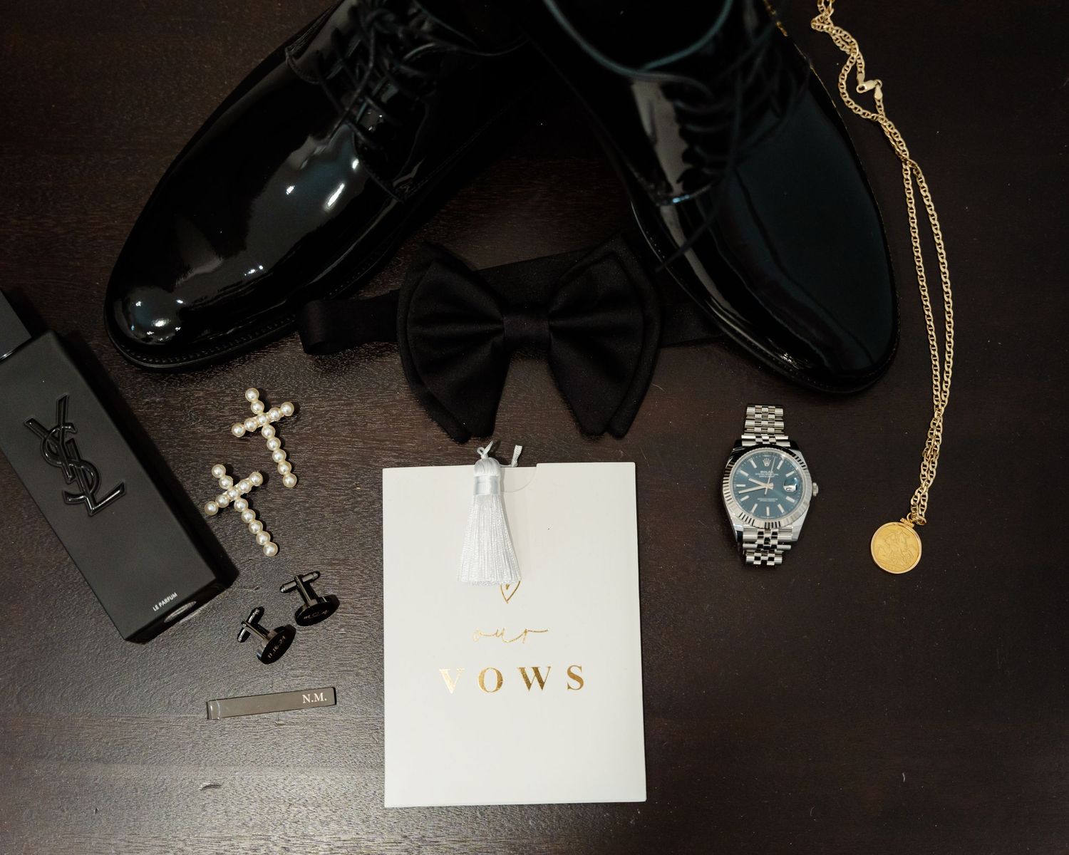Wedding accessories including bow tie, dress shoes, cross cufflinks, watch and gold necklace arranged on dark surface.