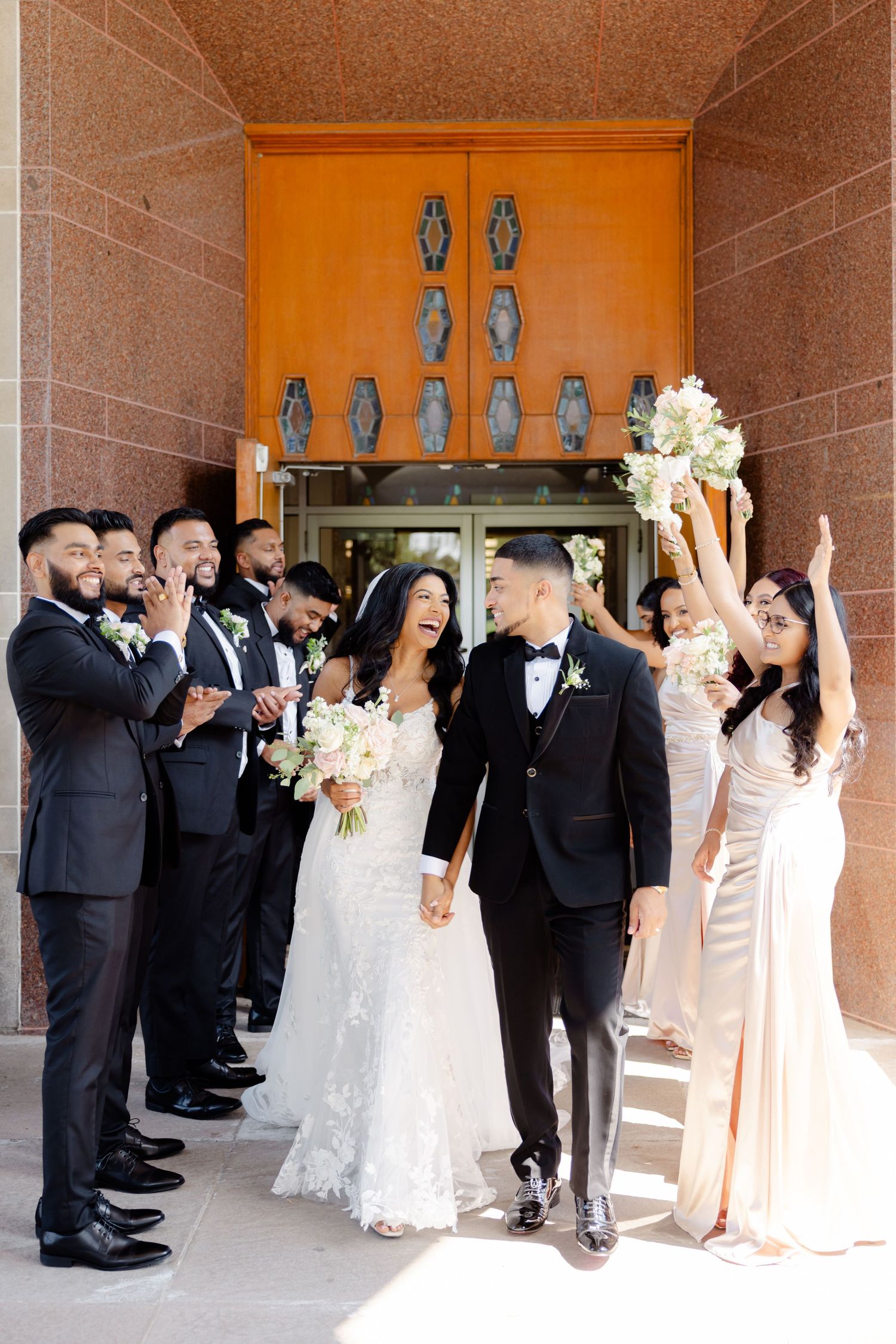 Wedding party celebrates outside ornate entrance with bridesmaids in blush dresses and groomsmen in black tuxedos.