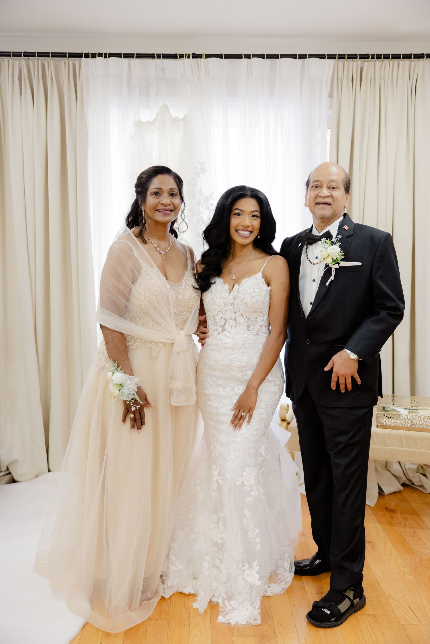 Three elegantly dressed wedding attendees pose together, with the bride in a white lace gown in the center.