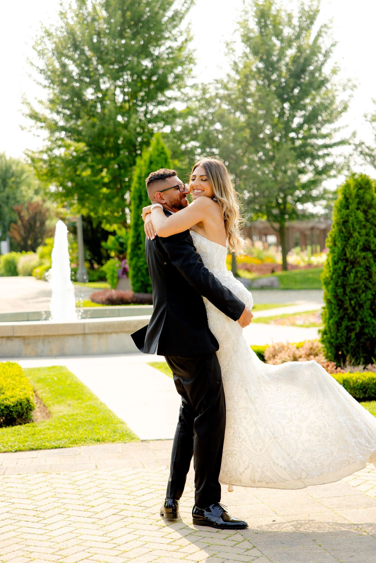 Newlyweds share intimate moment in sunlit garden with decorative fountain and cypress trees.