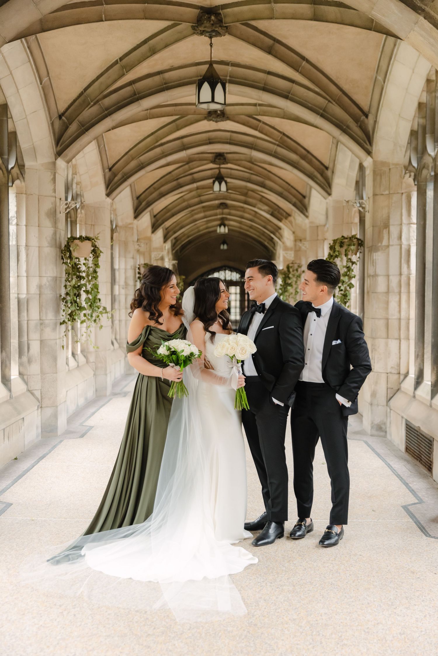 Wedding group shares candid moment in elegant stone hallway with hanging greenery.