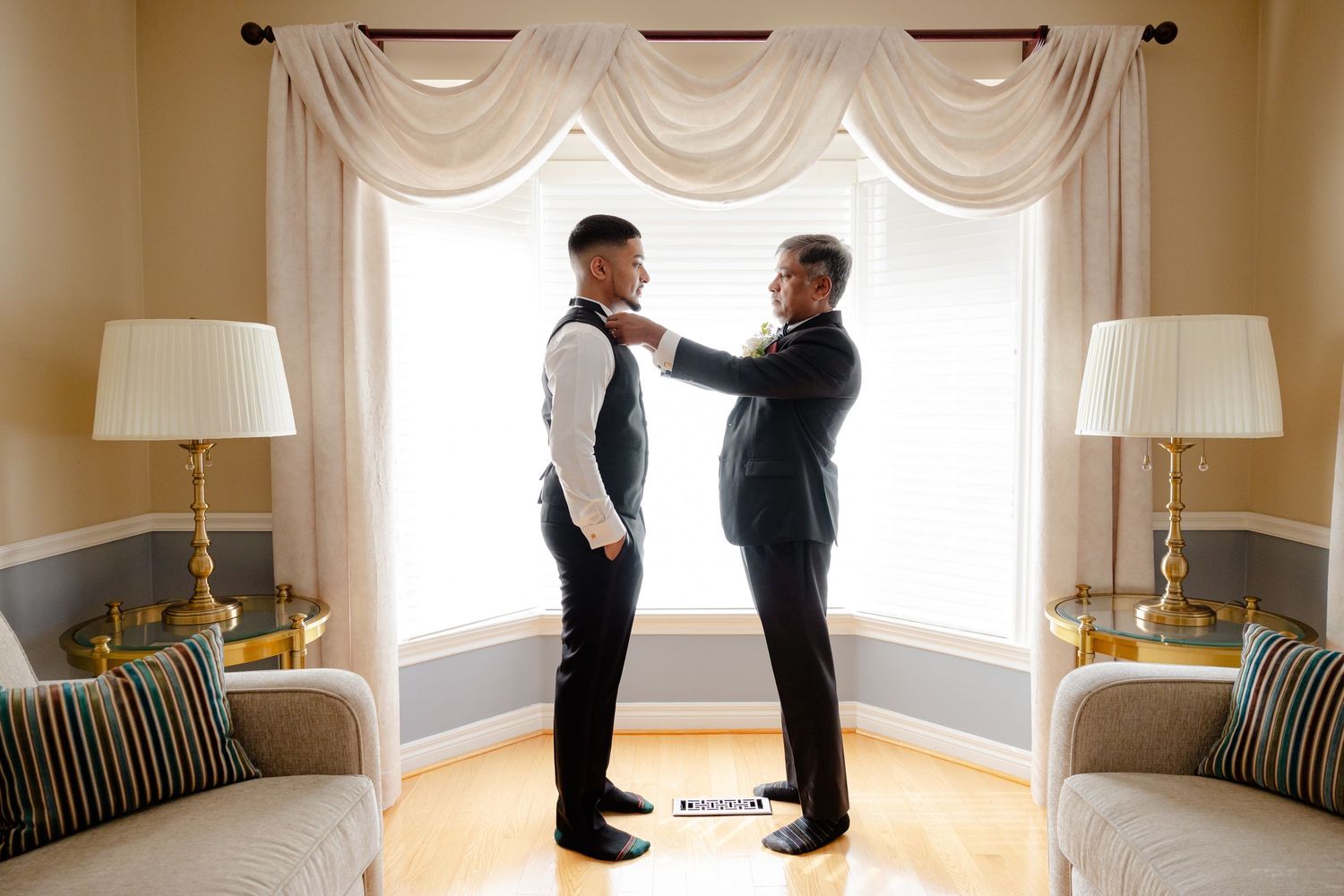 Two people in formal attire stand in a well-decorated room with elegant curtains and table lamps.