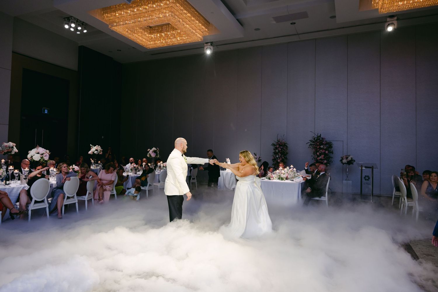 Wedding couple dancing in a misty reception hall with guests seated at round tables.