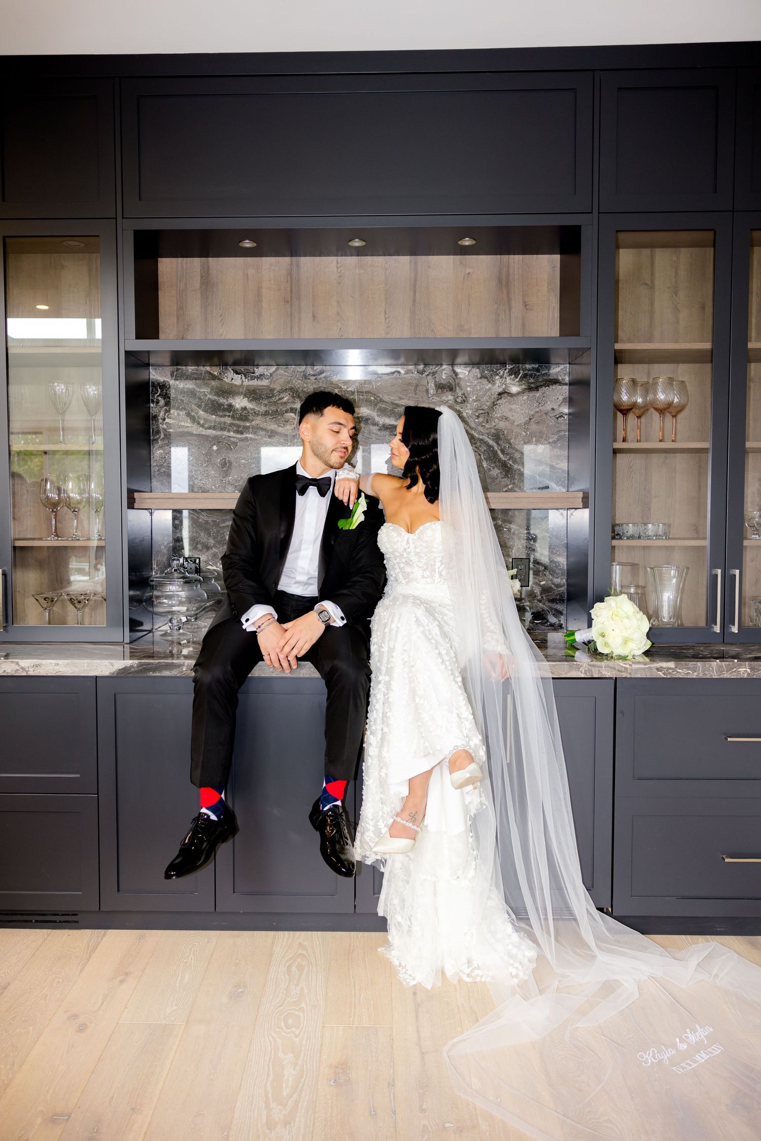 An elegant wedding photo of a couple posing in front of a dark kitchen with glass cabinets and marble backsplash.