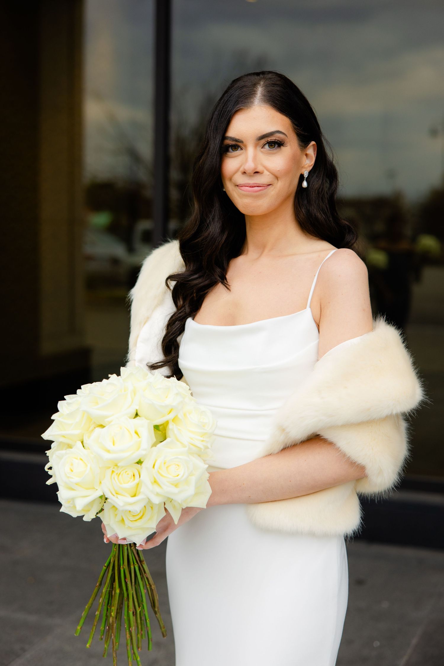 Bride in elegant white gown with fur wrap holds cream rose bouquet and smiles.