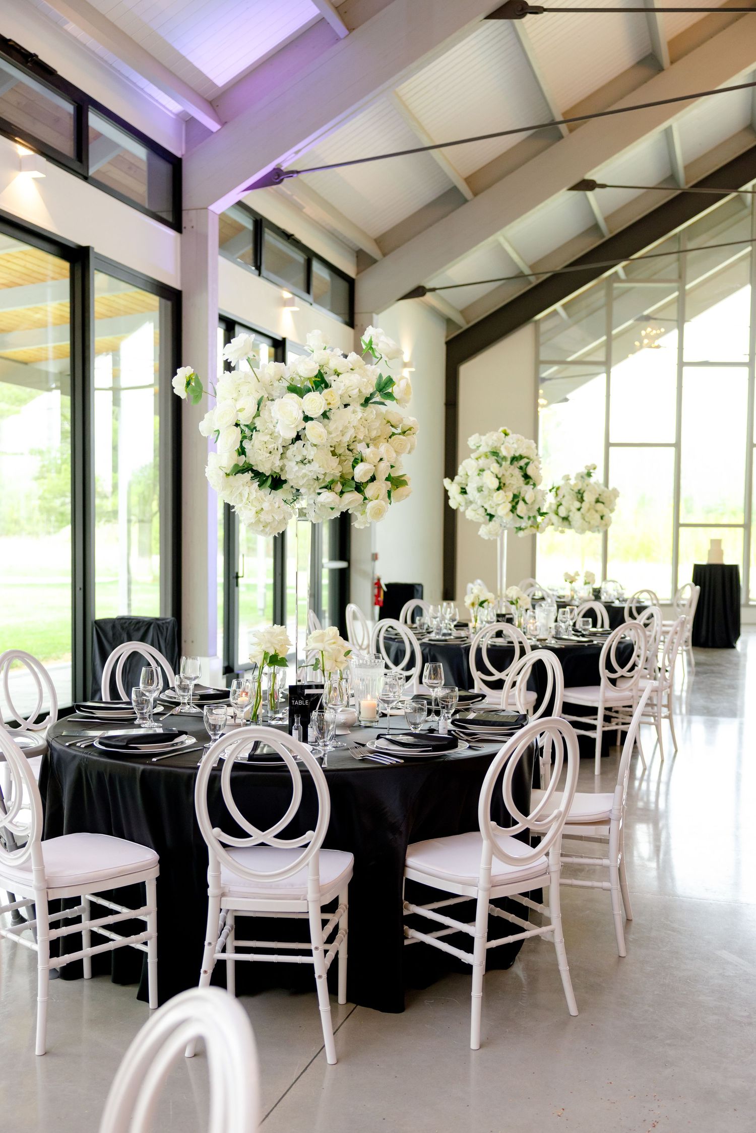A modern wedding reception setup with black tablecloths, white chairs and tall floral centerpieces.