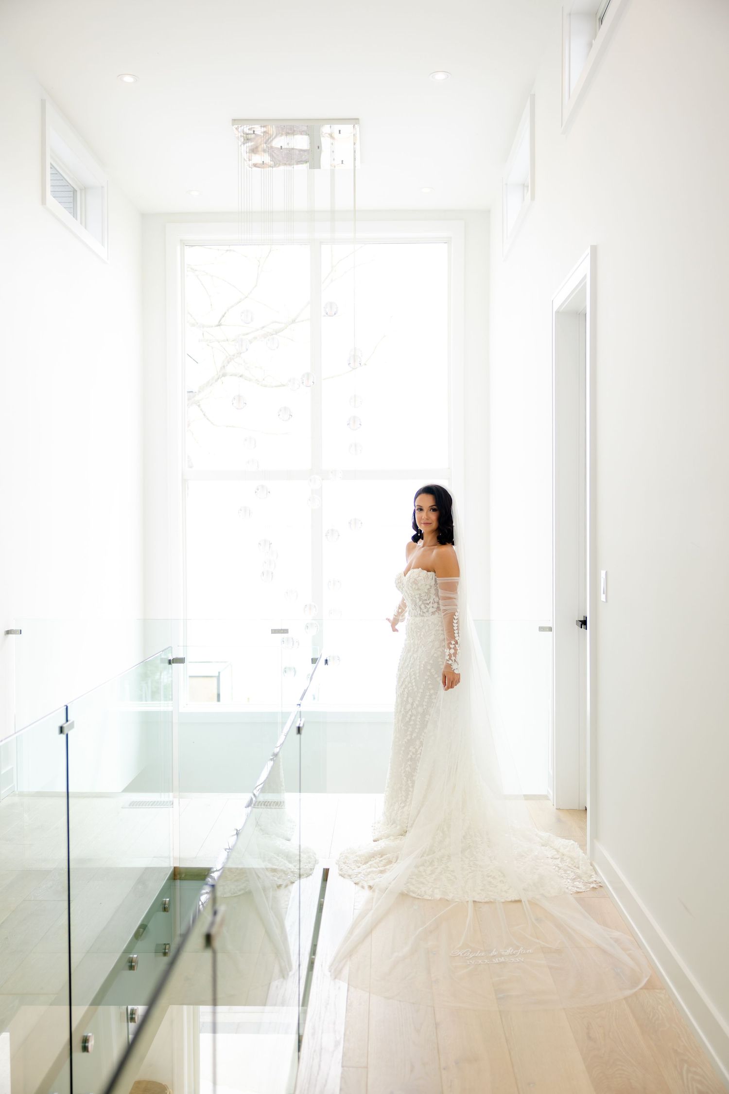An elegant bridal portrait in a bright white hallway featuring a fitted lace wedding dress with long sleeves.