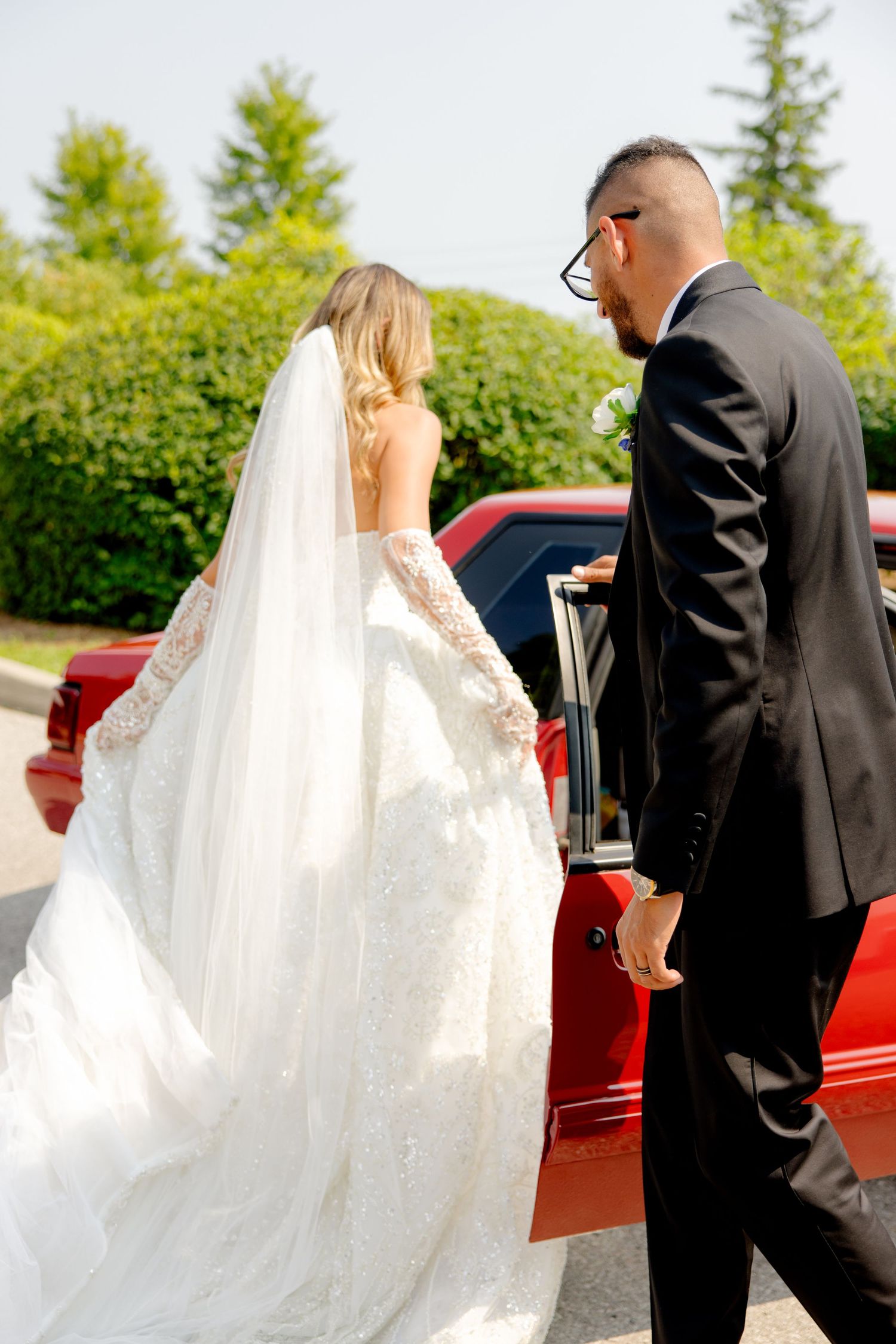 Elegant wedding couple standing by a red vintage car with beautiful greenery in the background.