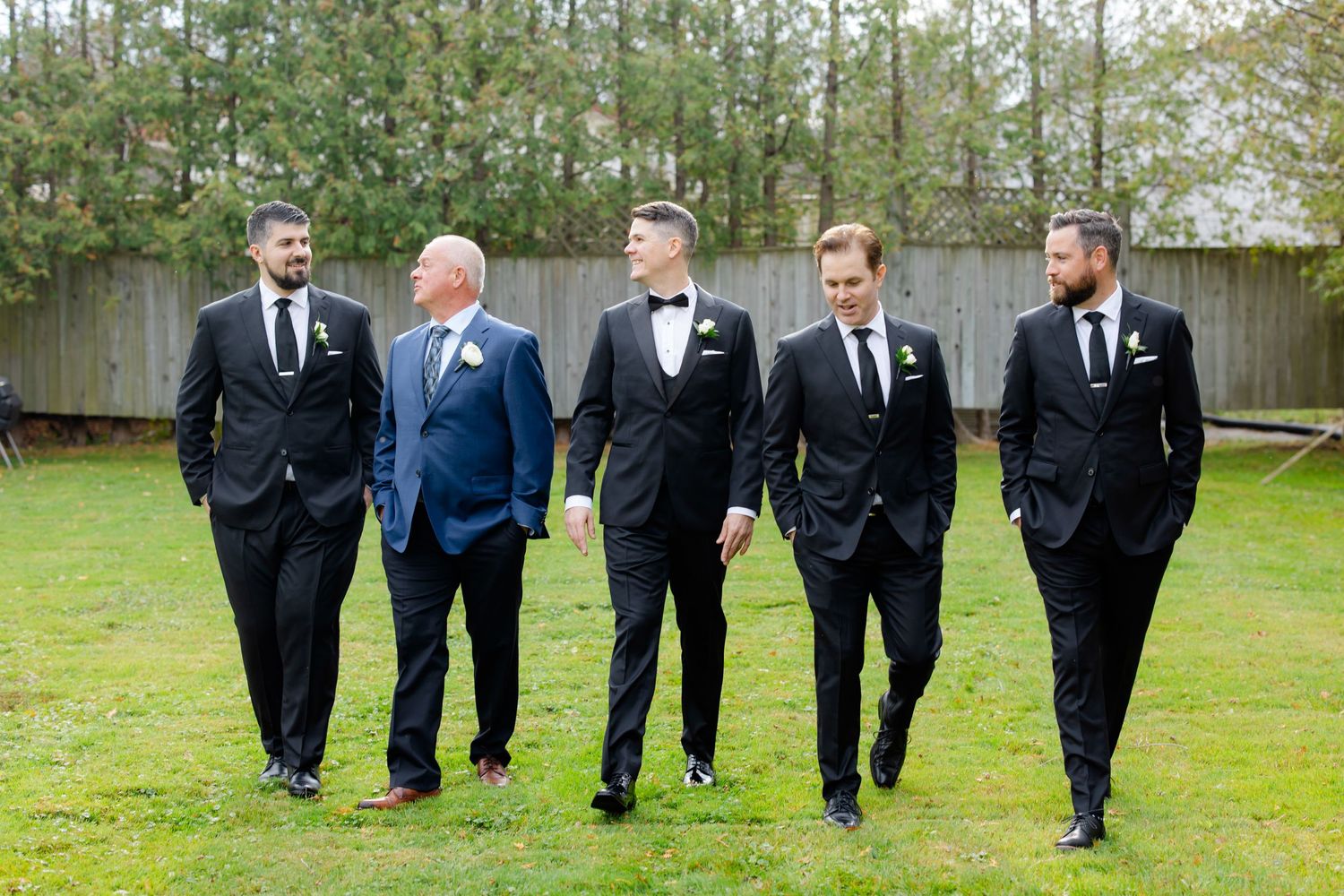 Five people in formal attire walking together on grass with trees in the background.