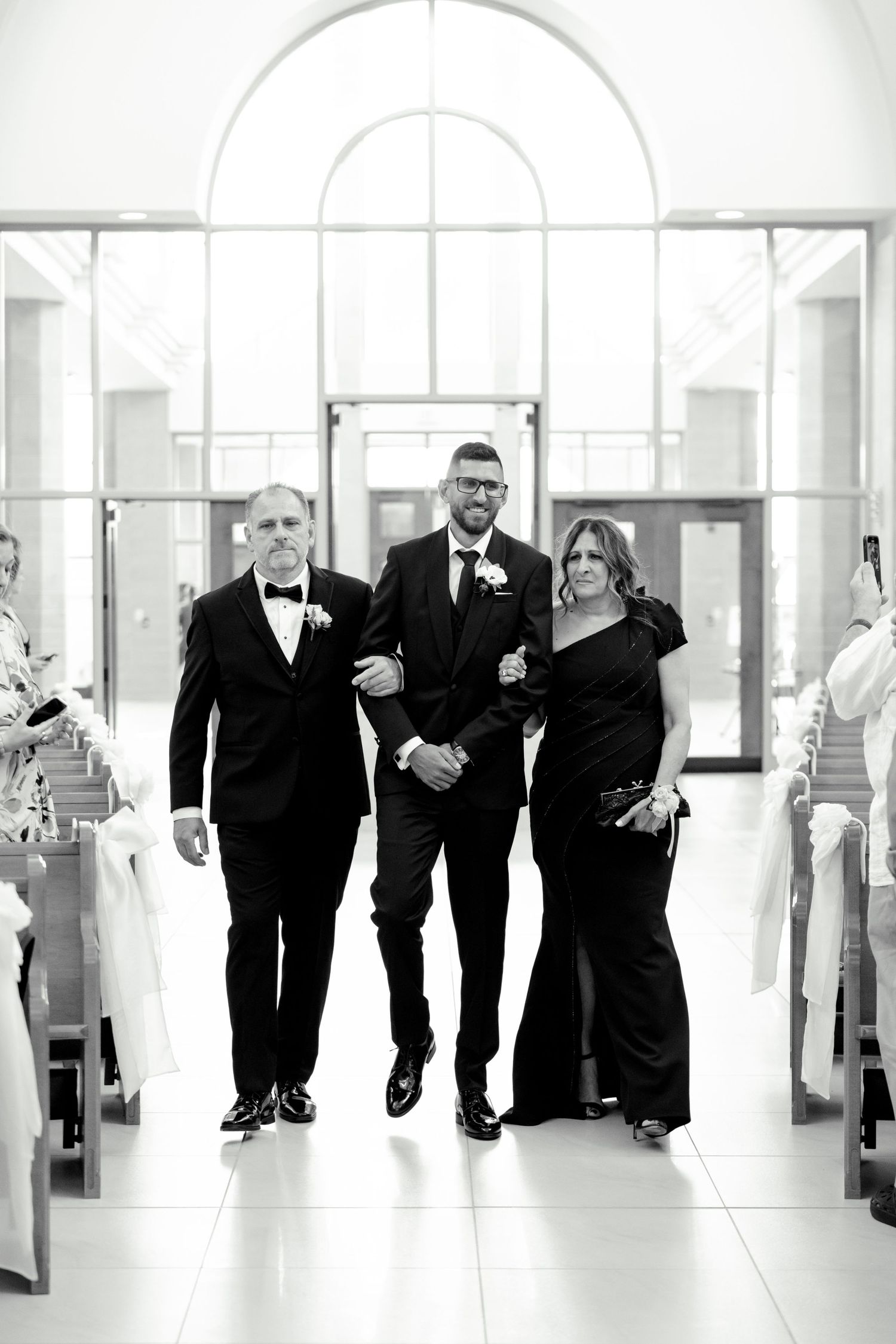 A black and white photo of three people walking down a bright church aisle.