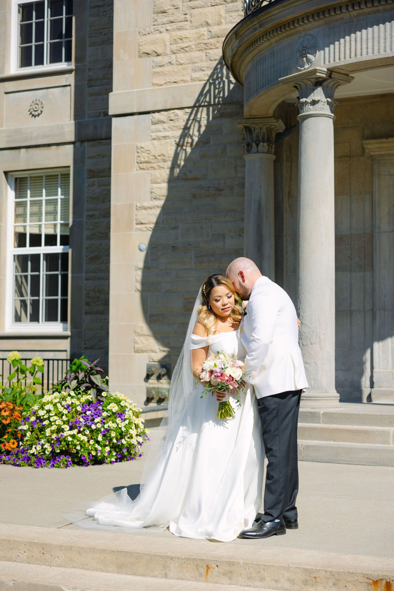 Wedding couple embracing on stone steps beside colorful flower garden and classical columns.