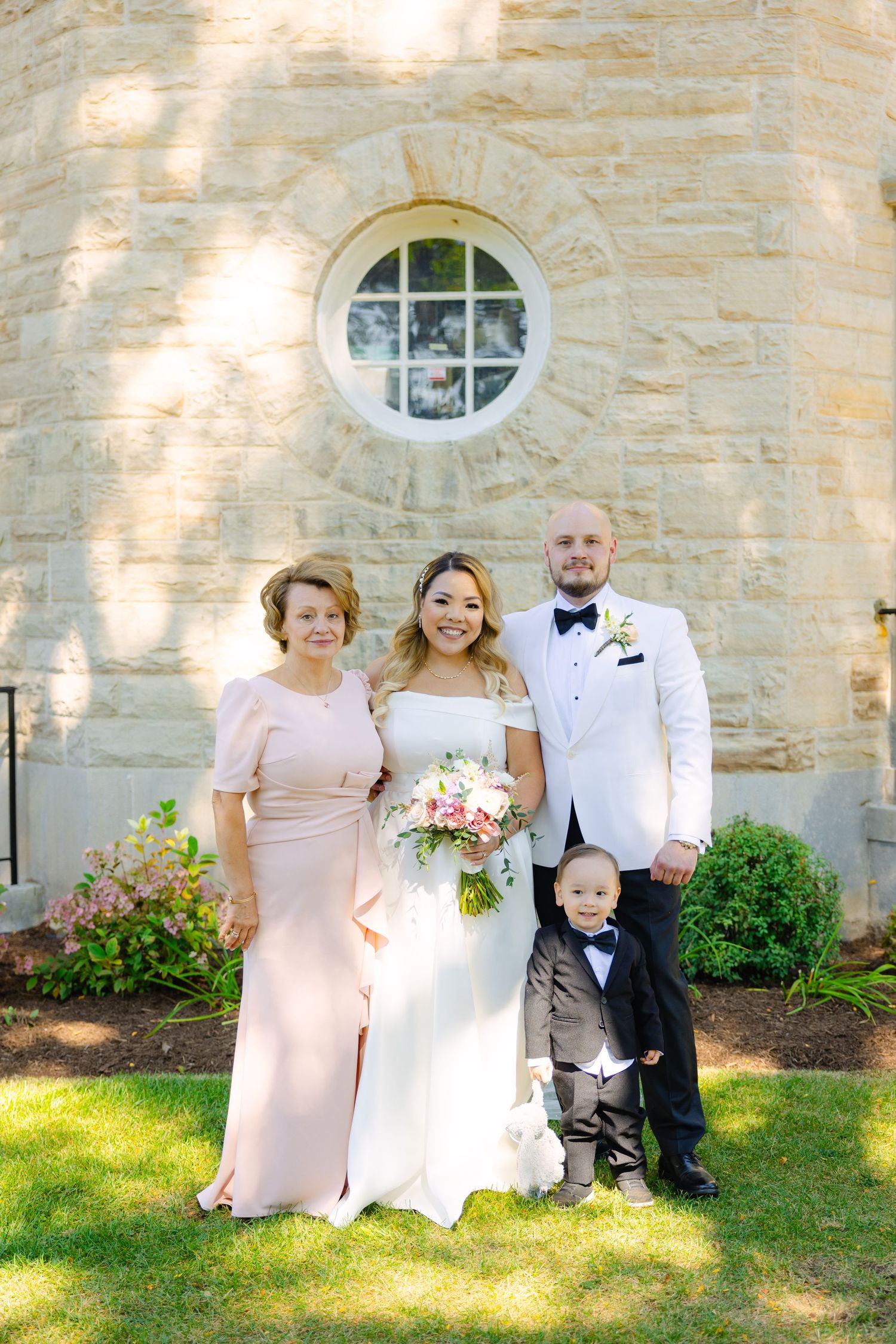Small wedding group with young child poses in formal attire against stone wall.