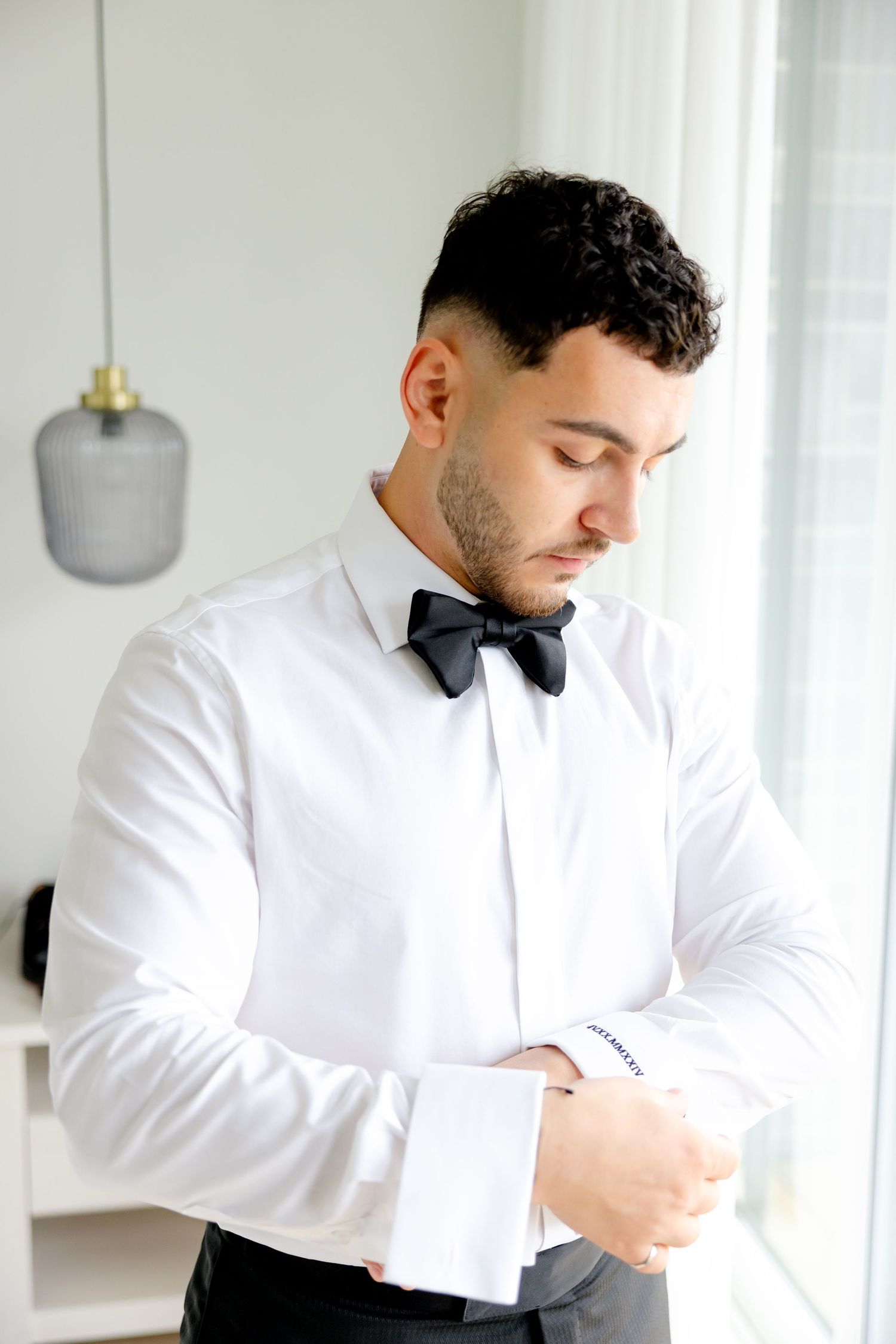 Person in white dress shirt and bow tie adjusts cuff by window in formal attire.