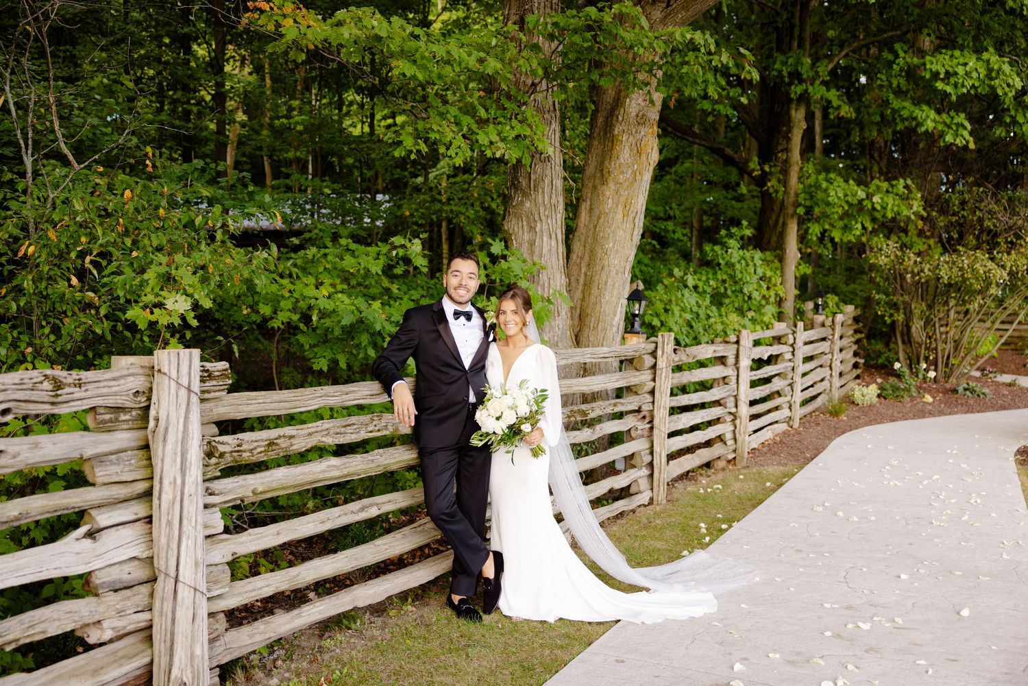 A couple in wedding attire poses along a rustic fence with trees in the background.