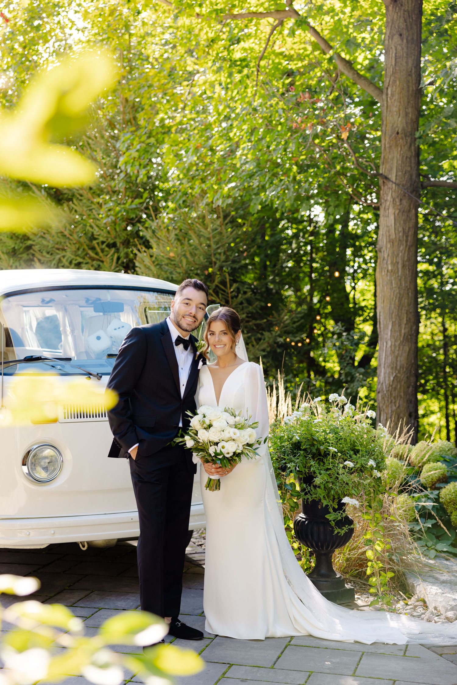 Newlyweds stand next to a classic white vehicle surrounded by lush garden landscaping.