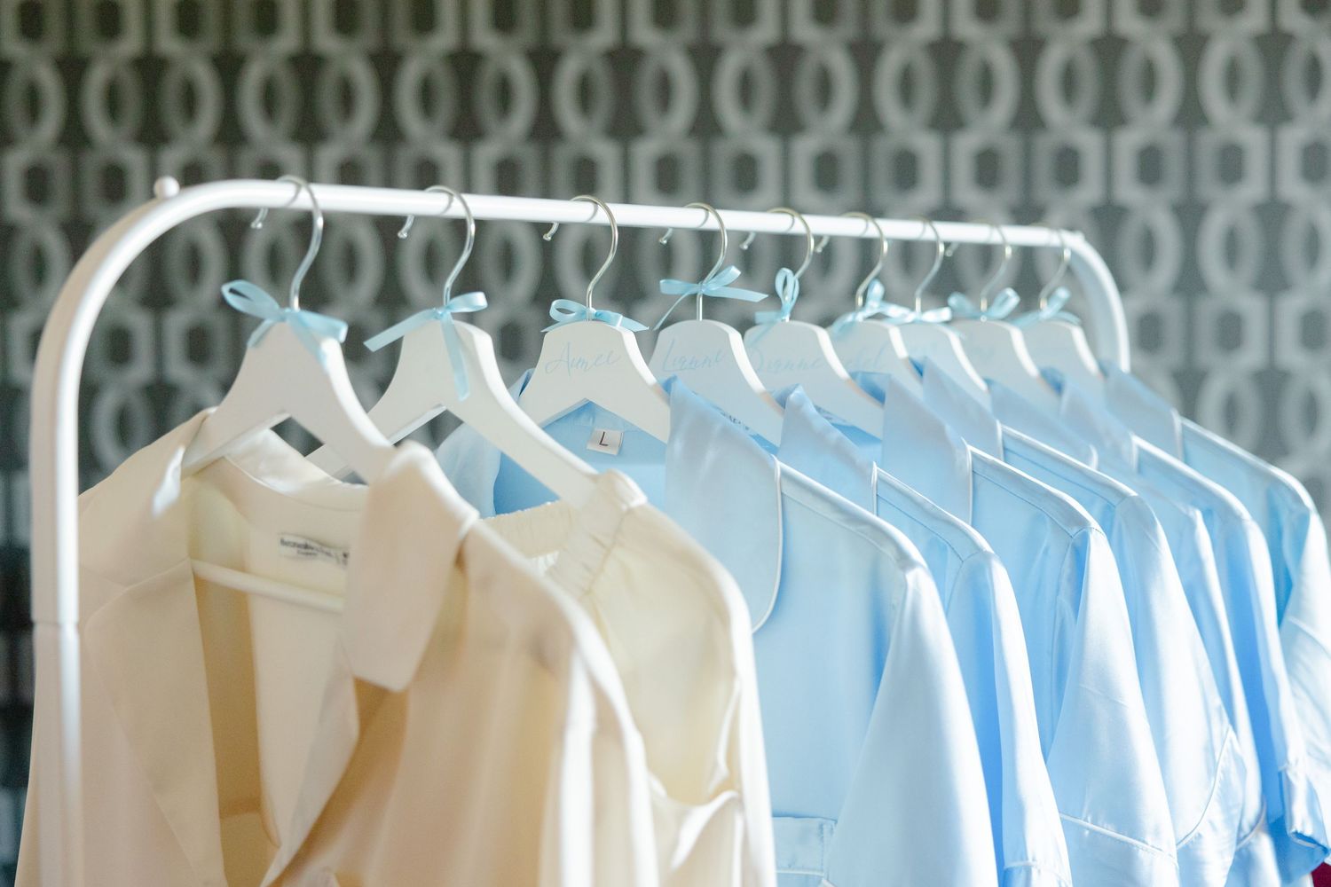 White and light blue dress shirts hanging neatly on clothing rack against patterned wallpaper.